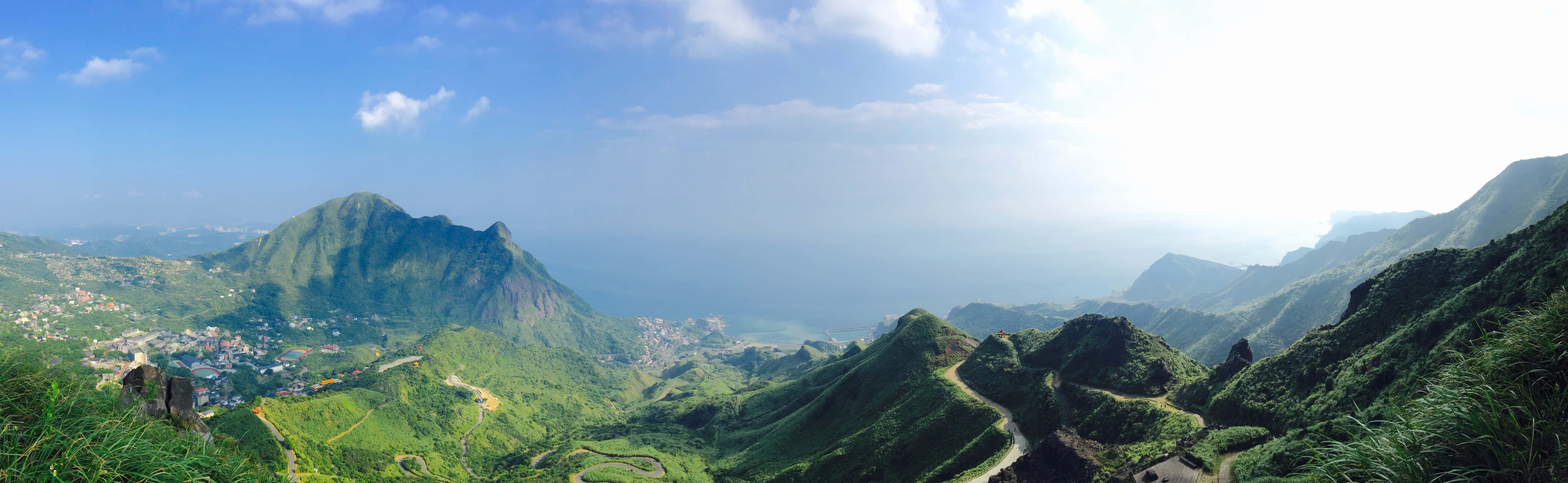山景超高清雙螢幕壁紙 壁紙 — 翠綠山巒的全景，蜿蜒的道路和沿海村莊，在明亮的藍天下有零星雲朵。