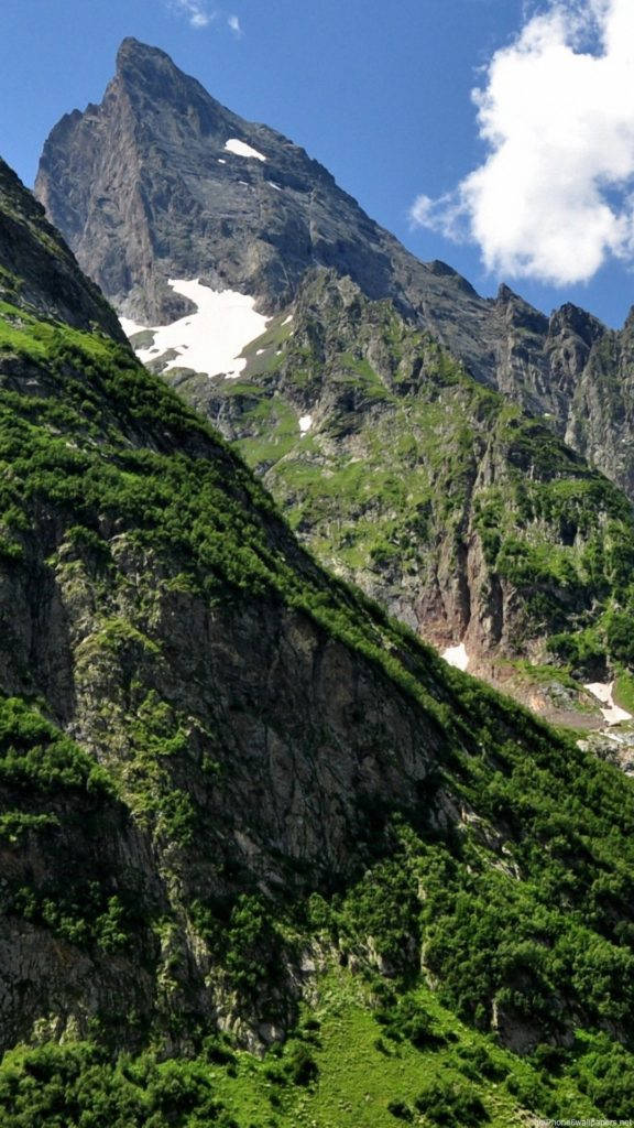 山林自然 iPhone 背景 壁紙 — 高聳的岩石山峰，帶有雪斑，在明亮的藍天下有蓬鬆的白雲。