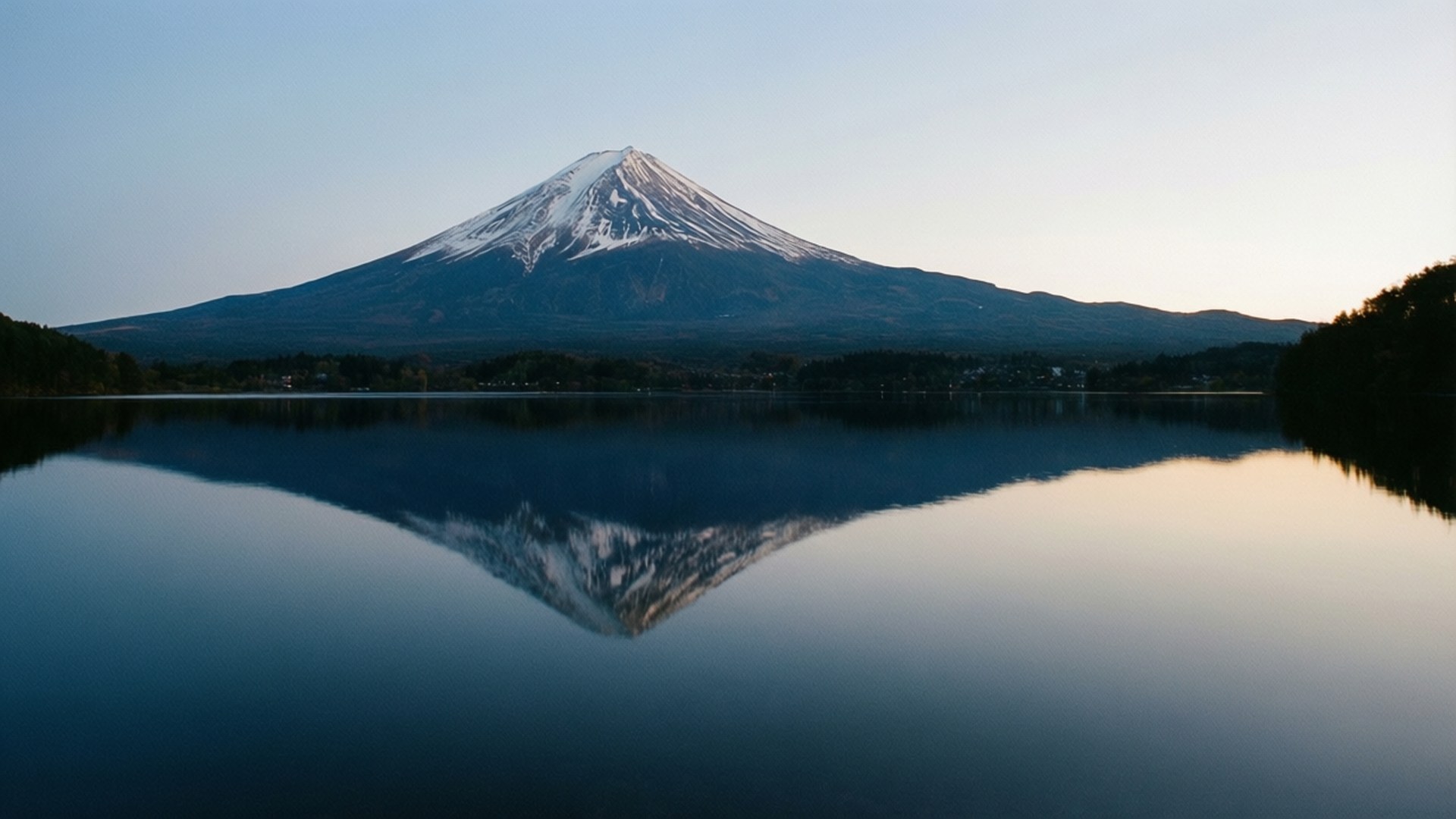 山湖倒影 壁紙 — 白雪覆蓋的富士山在黎明時分完美倒映在平靜的湖面上，呈現柔和的藍色和金色色調。