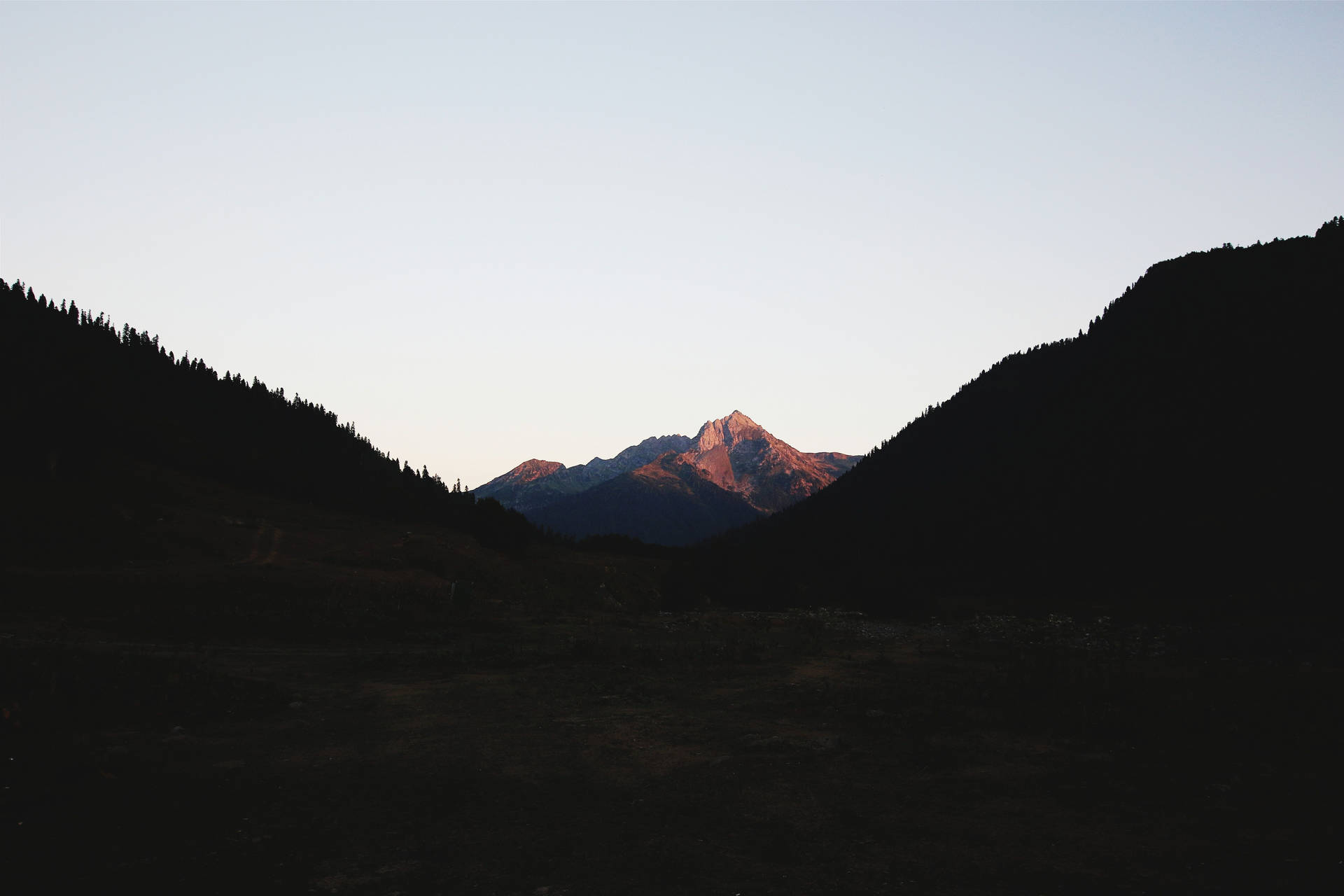 一座山峰沐浴在日落光芒中，被黑暗的森林山丘環繞。