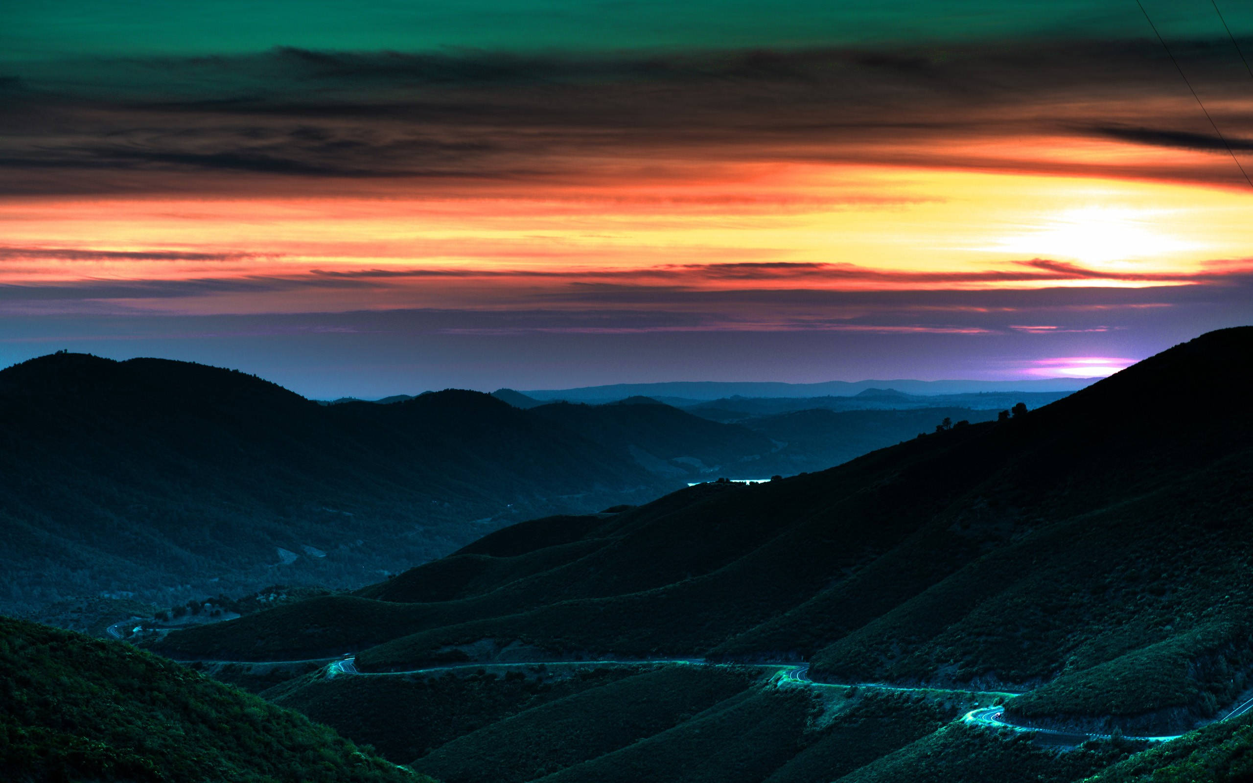 山腰夕照 壁紙 — 一條蜿蜒的道路在火紅的夕陽天空下蜿蜒穿過黑暗起伏的山丘，天空佈滿橙色、紫色和藍綠色的條紋。