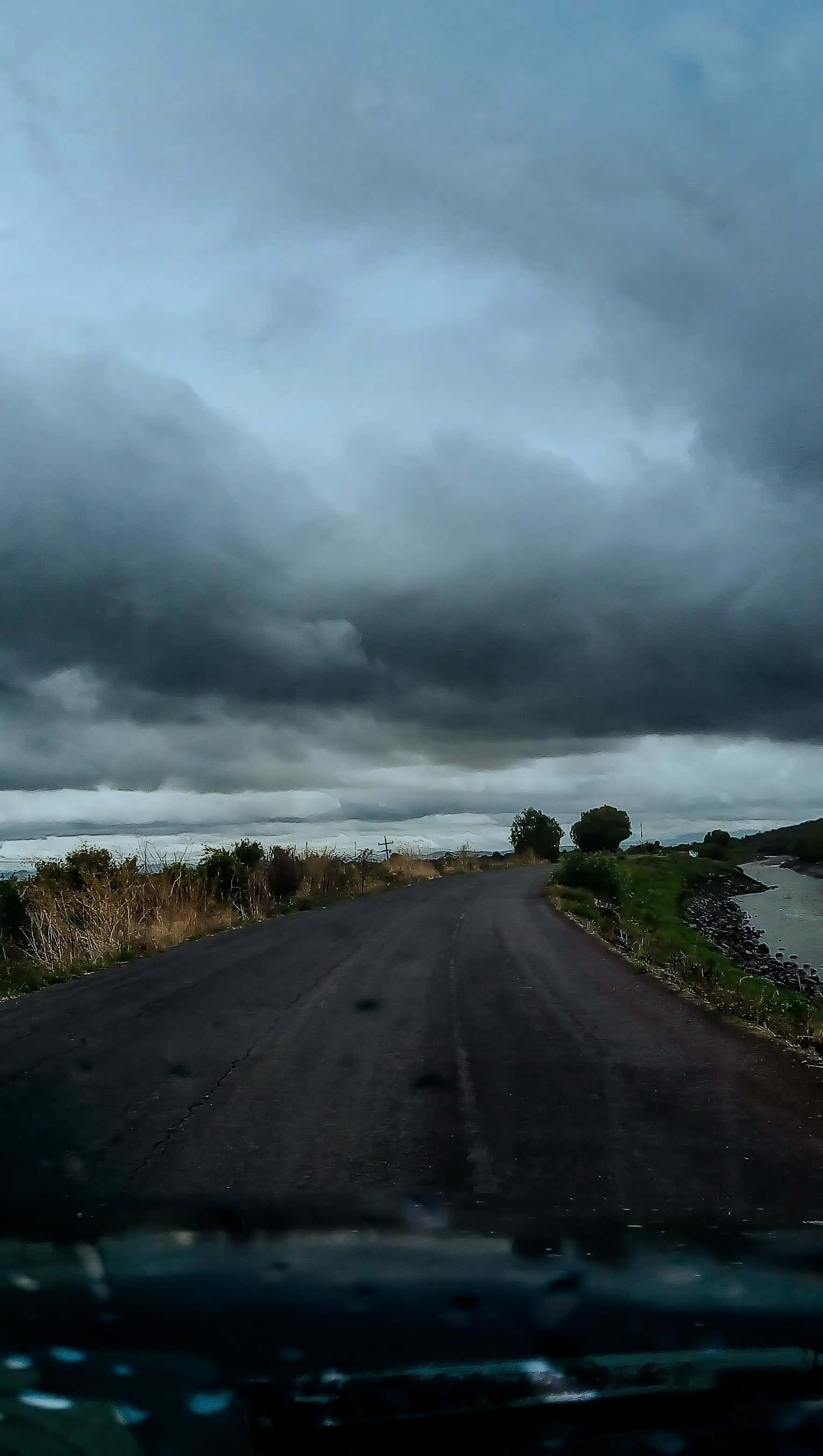 山間晨雨景象 壁紙 — 一條被雨水打濕的道路蜿蜒伸向暴風雨的地平線，烏雲籠罩著遠處的水域。