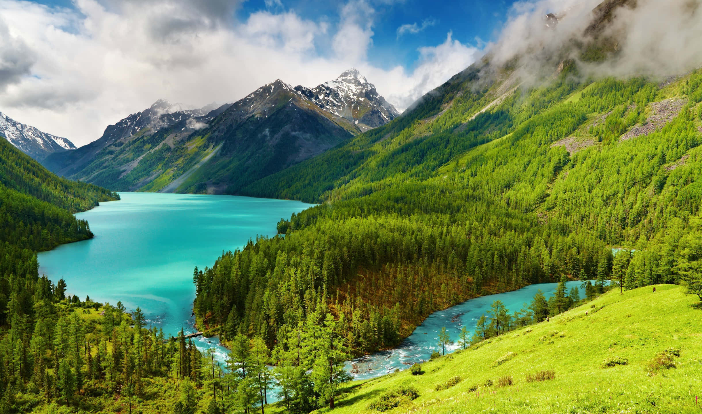 山間湖泊與樹木山巒 壁紙 — 一個充滿活力的藍綠色湖泊，坐落在鬱鬱蔥蔥的綠色山脈中，山頂積雪，天空多雲，藍天白雲。