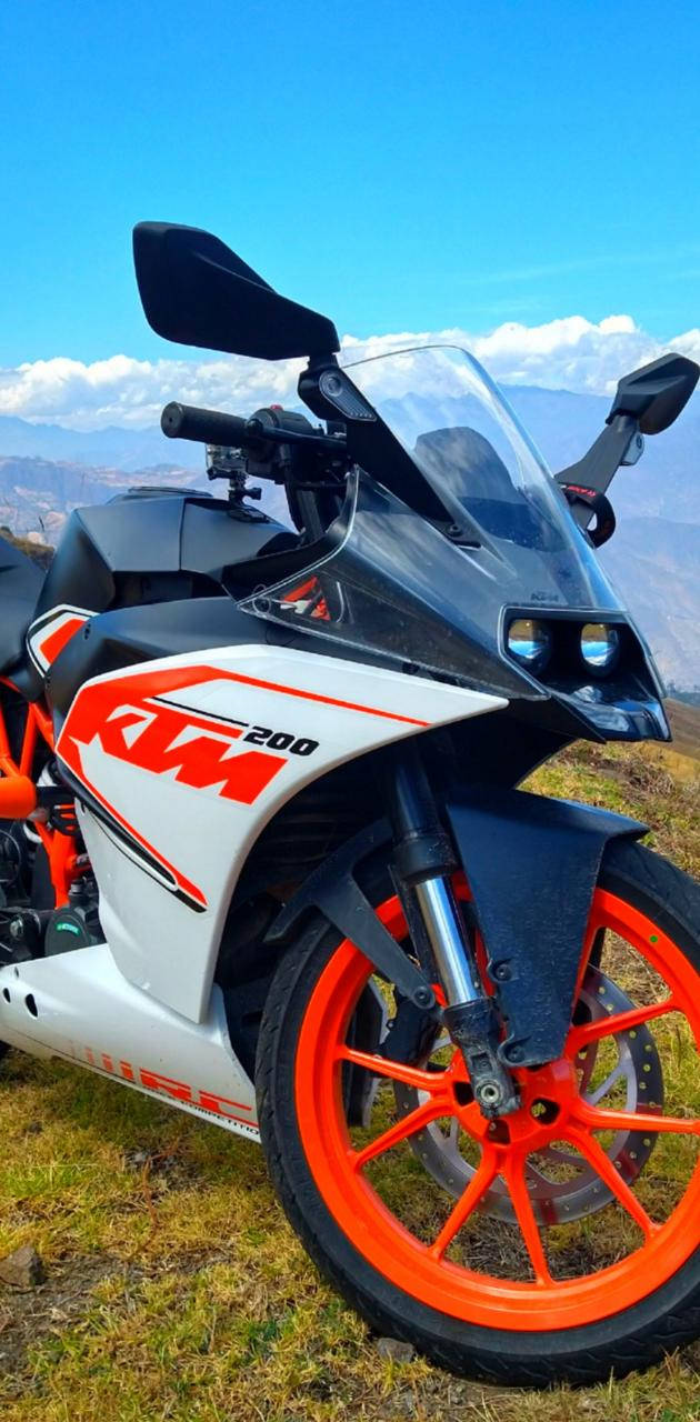 山頂上的KTM RC 200 壁紙 — 一架白色和橙色的KTM 200電單車停泊在草地上，背景有山脈和藍天。