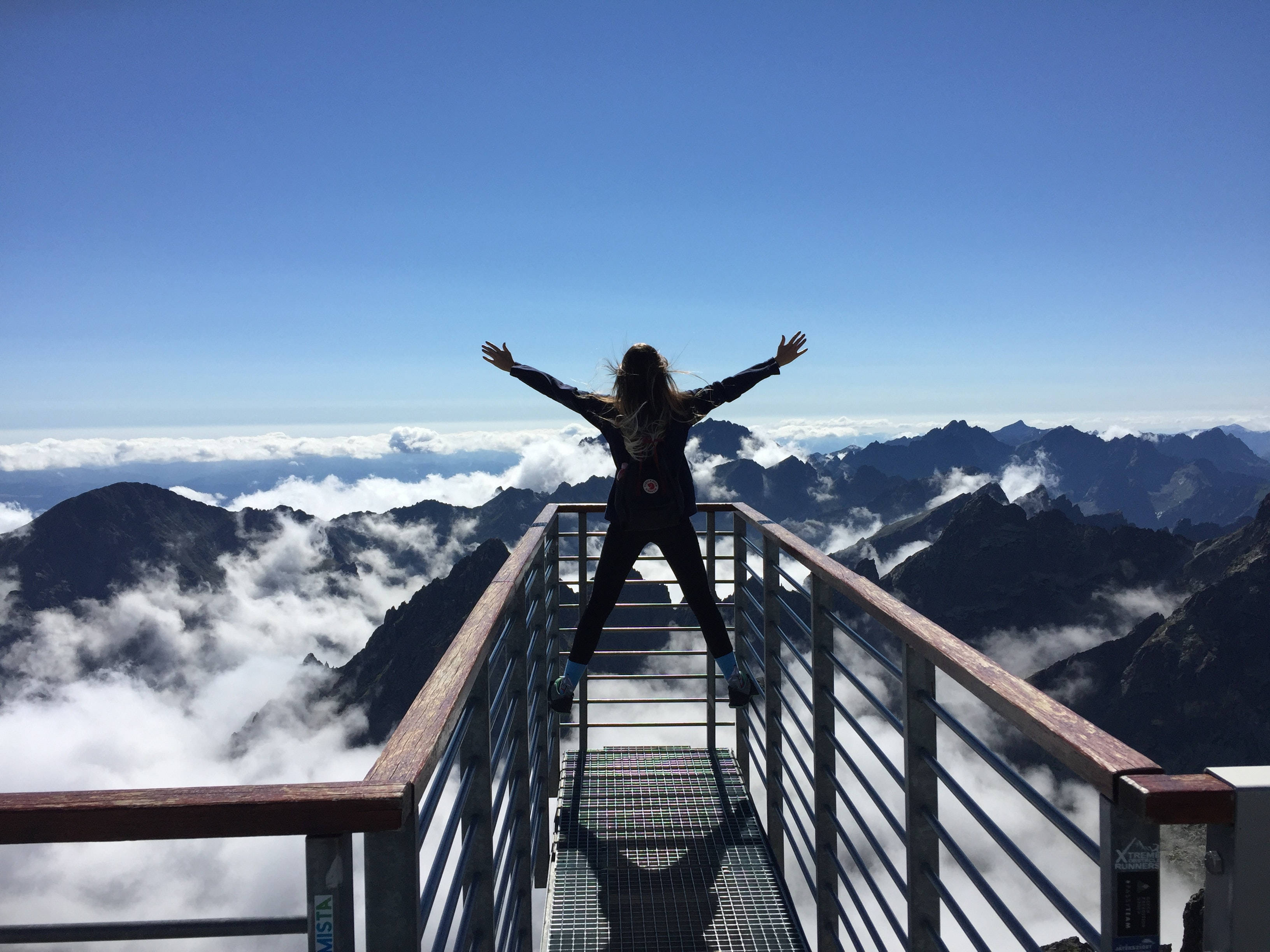 山頂度假 壁紙 — 一個人站在山頂觀景台上，雙臂張開，擁抱廣闊的雲海和下方崎嶇的山峰，頭頂是清澈的藍天。