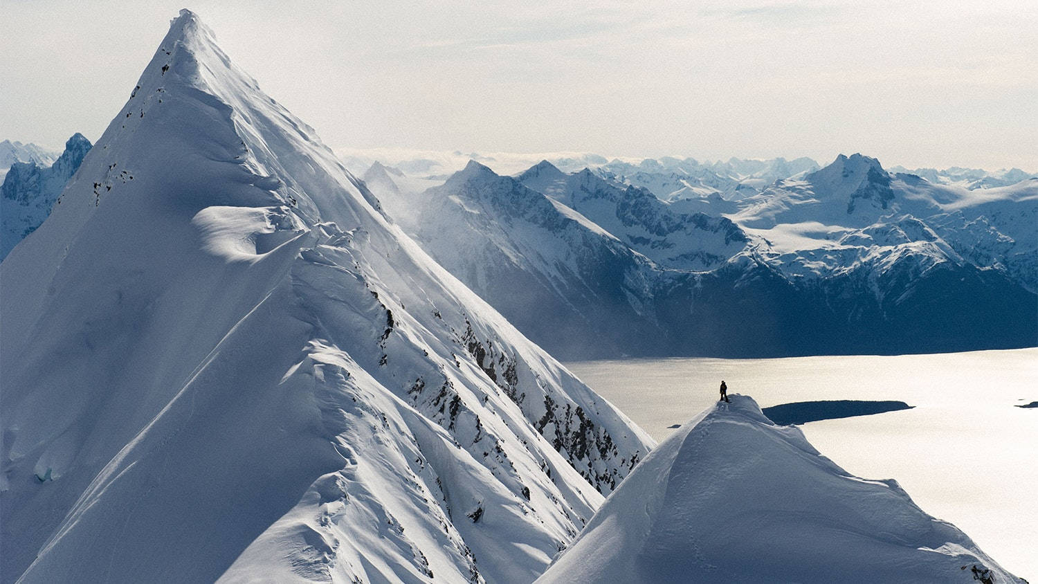 山頂滑雪 壁紙 — 一位孤獨的登山者站在白雪覆蓋的山脊頂端，俯瞰著廣闊的冰凍湖泊和遠處的山峰，天空蒼白。