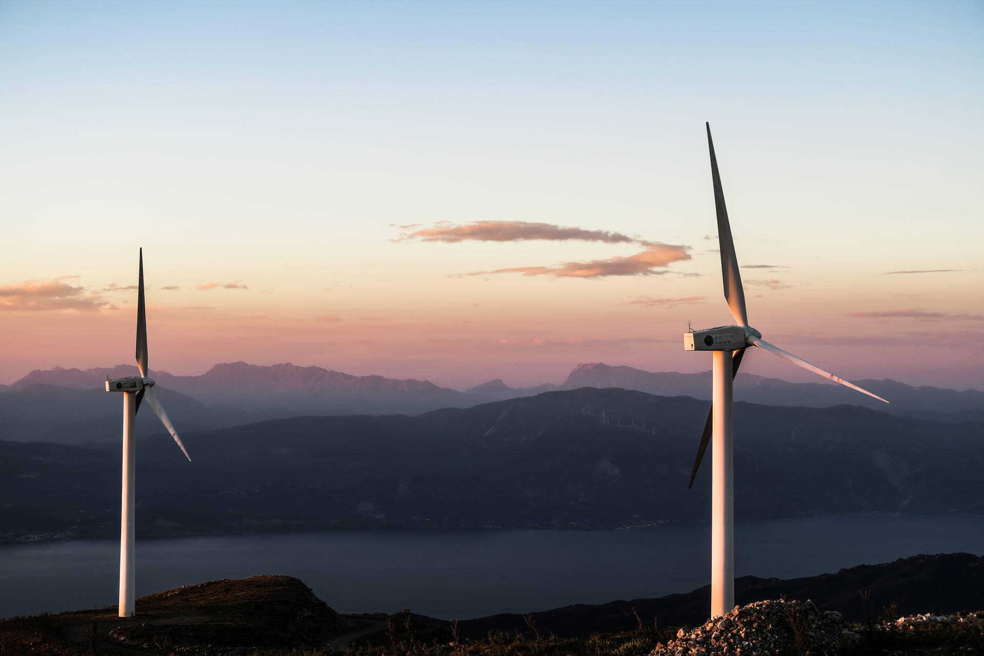 夕陽下，兩座風力發電機位於俯瞰山巒和海洋的山丘上。