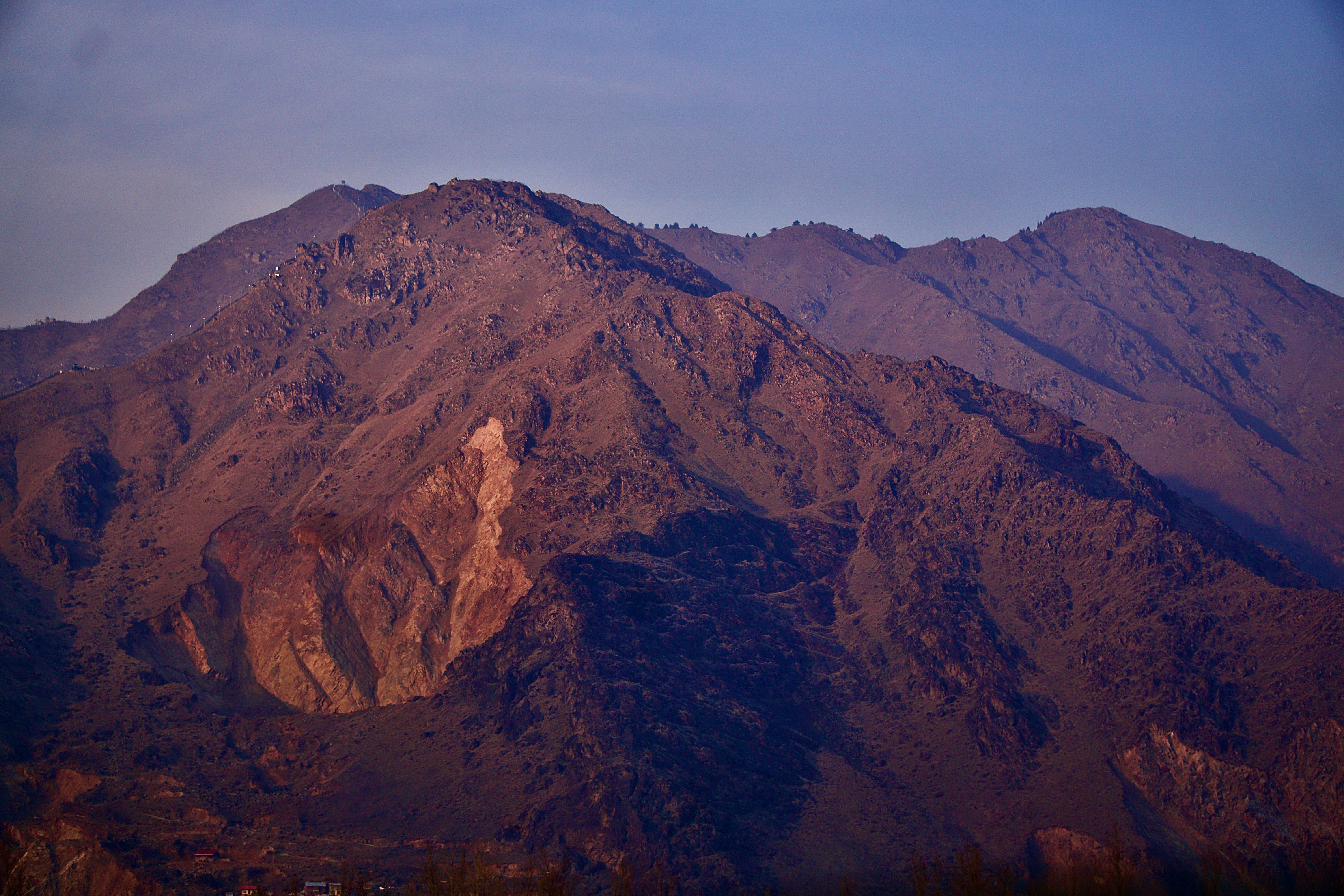 岩石山地地形 壁紙 — 崎嶇的山峰沐浴在溫暖的日落光芒中，在柔和的藍天下，光禿的山坡上投下長長的影子。