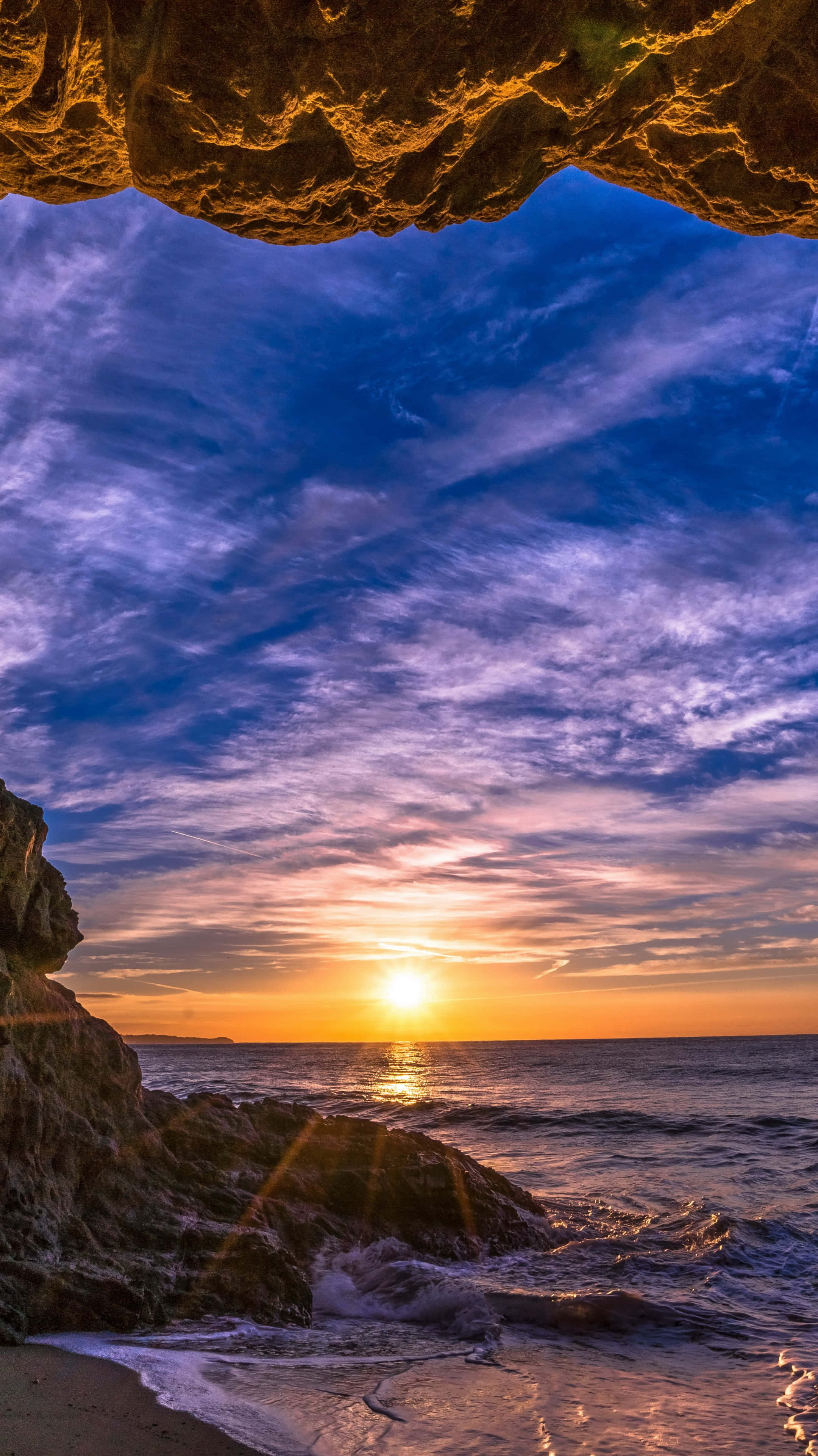 岩石洞穴視角 Android 馬利布背景 壁紙 — 夕陽透過岩石洞穴開口照耀，下方是洶湧的海洋，天空呈現鮮豔的紫色和橙色。