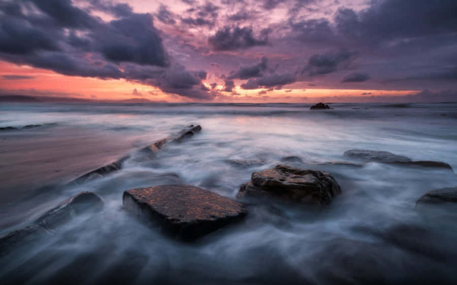 岩石與水畔的美麗日落 壁紙 — 日落時分的長時間曝光海景，平滑的水流在深色岩石周圍流動，天空呈現戲劇性的紫色和橙色。
