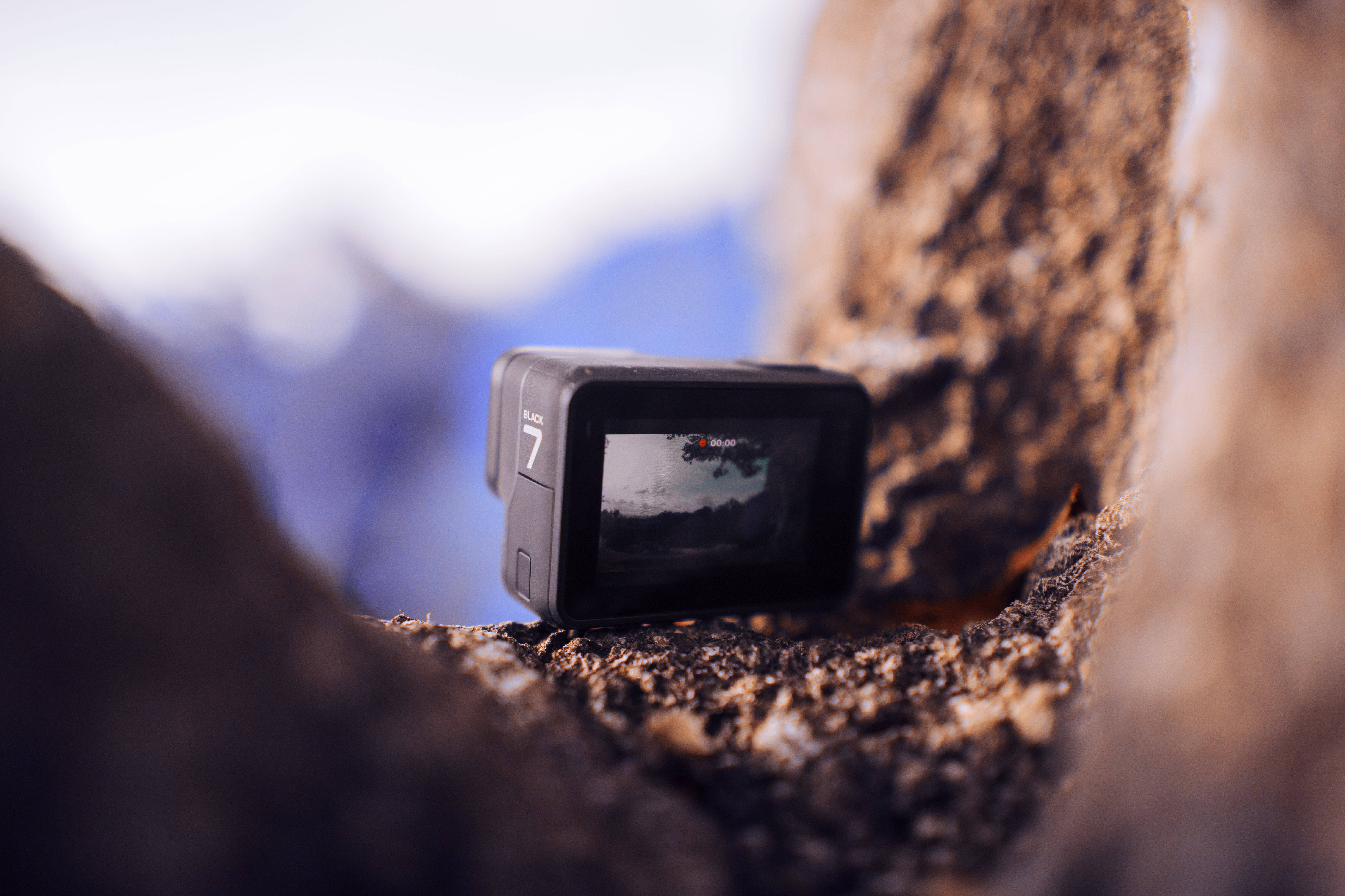 岩石邊緣上的GoPro 壁紙 — 一部GoPro 7相機嵌在岩石縫中，屏幕上捕捉到模糊的山景。