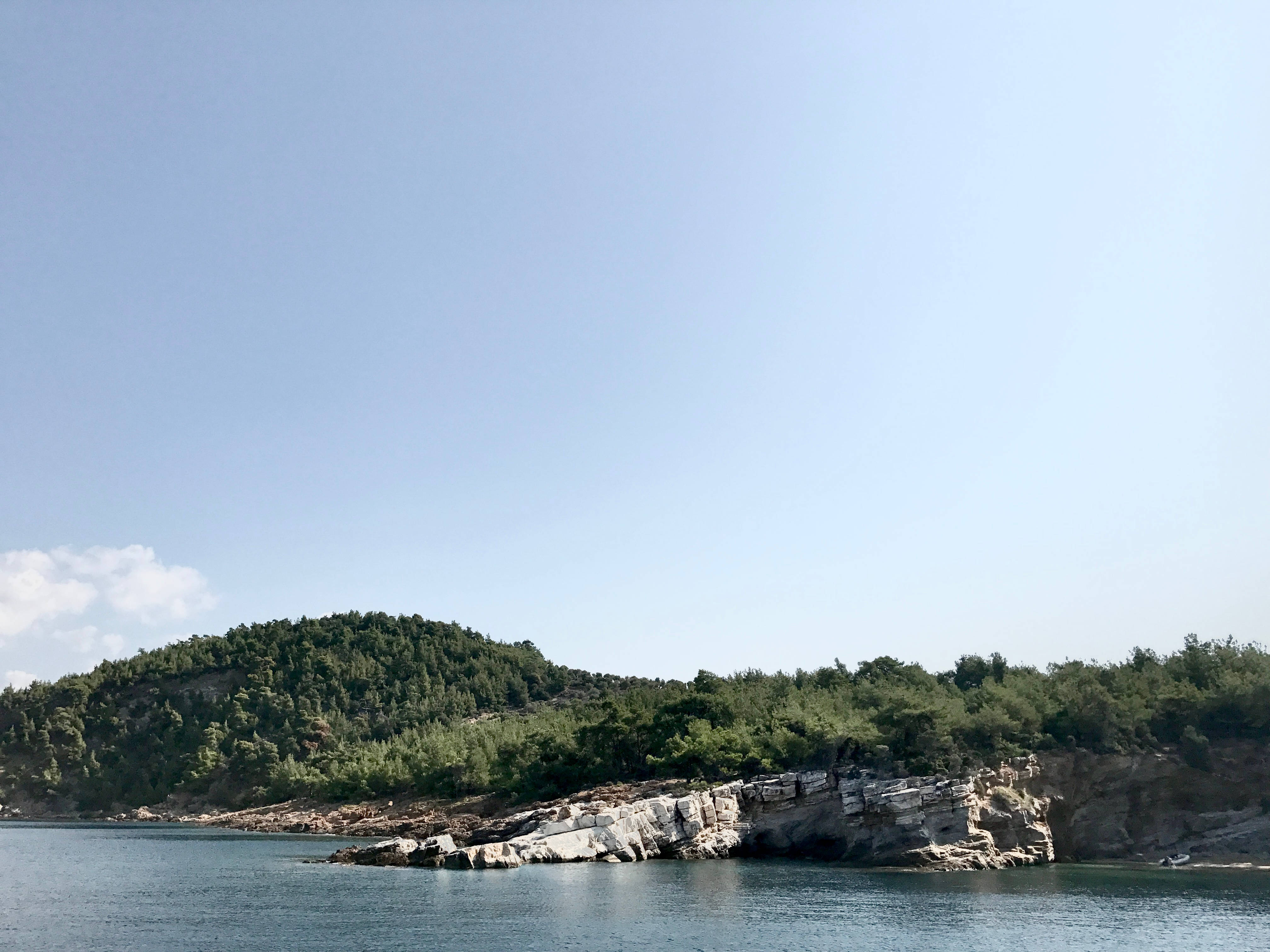 島嶼意境筆電壁紙 壁紙 — 平靜的藍色湖泊與岩石海岸線接壤，岸邊有茂密的綠樹，天空晴朗。