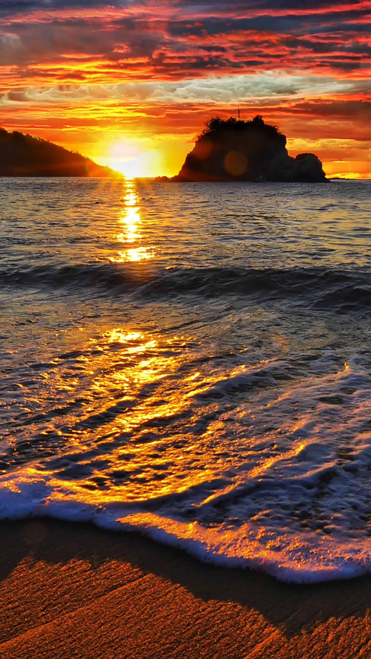 島嶼海灘的日落 壁紙 — 熾熱的夕陽將海洋染成金色，海浪拍打著沙灘，旁邊是岩石島的剪影。