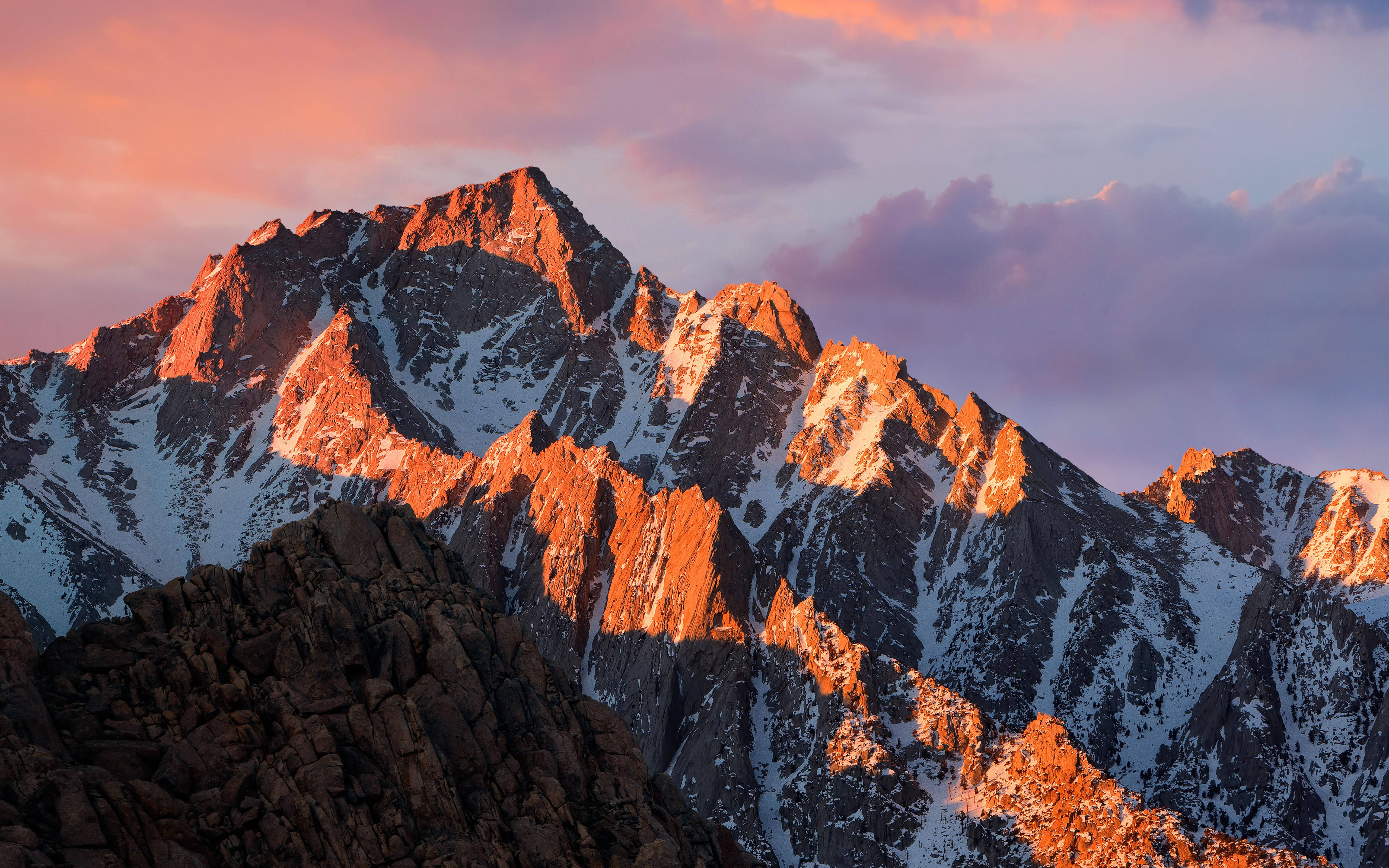 崎嶇山巒 MacBook Pro 4K 壁紙 — 夕陽將崎嶇的積雪山峰沐浴在溫暖的橙色光芒中，映襯著柔和的粉紅色和紫色天空。