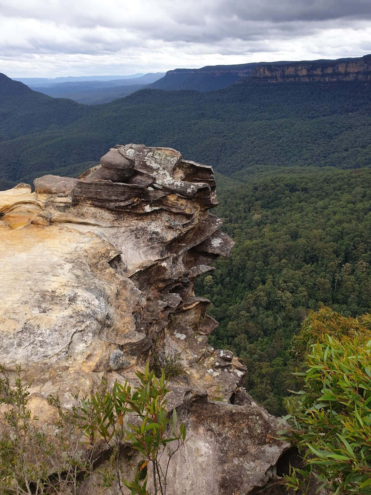 Blue Mountains National Park 桌布 壁紙 — 崎嶇的懸崖邊緣俯瞰著廣闊的綠色山谷，天空多雲，遠處有層層疊疊的山脈。