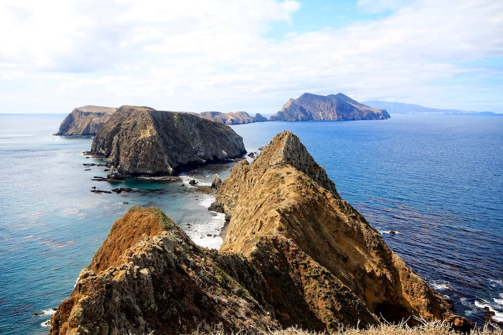 California Island 桌布 壁紙 — 崎嶇的海岸懸崖在部分多雲的天空下墜入深藍色的海洋，地平線上有遙遠的島嶼。