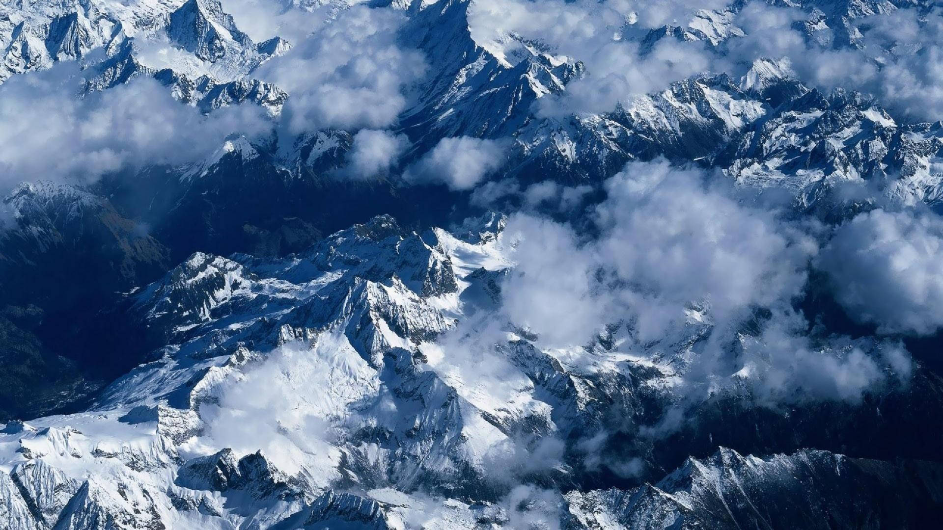 崑崙雪山 壁紙 — 鳥瞰圖顯示鋸齒狀的雪峰在明亮的日光下刺穿旋轉的白雲。