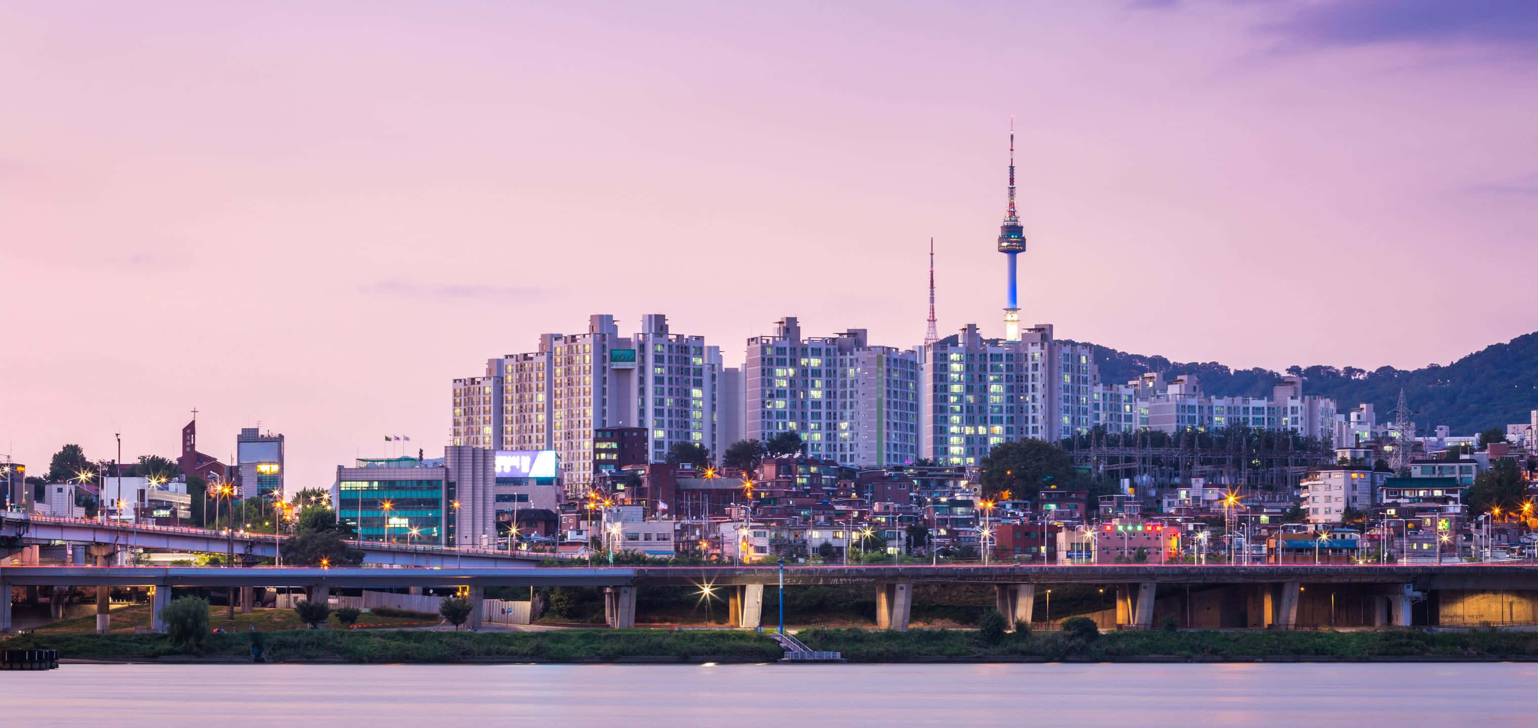 巍峨山脈與如畫天空 壁紙 — 寧靜的暮色城市景觀，河流反射橋樑和建築物的燈光，在柔和的紫色天空下。