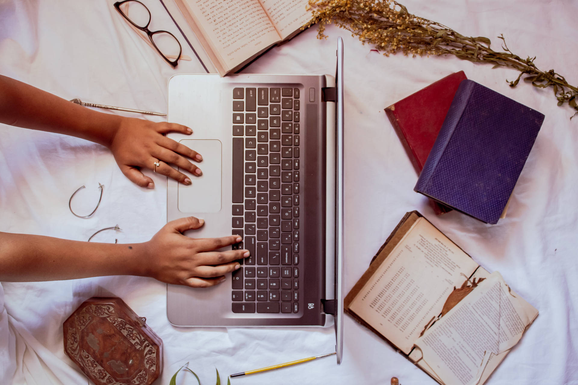 Hands typing on a laptop surrounded by books and dried flowers on a bed.