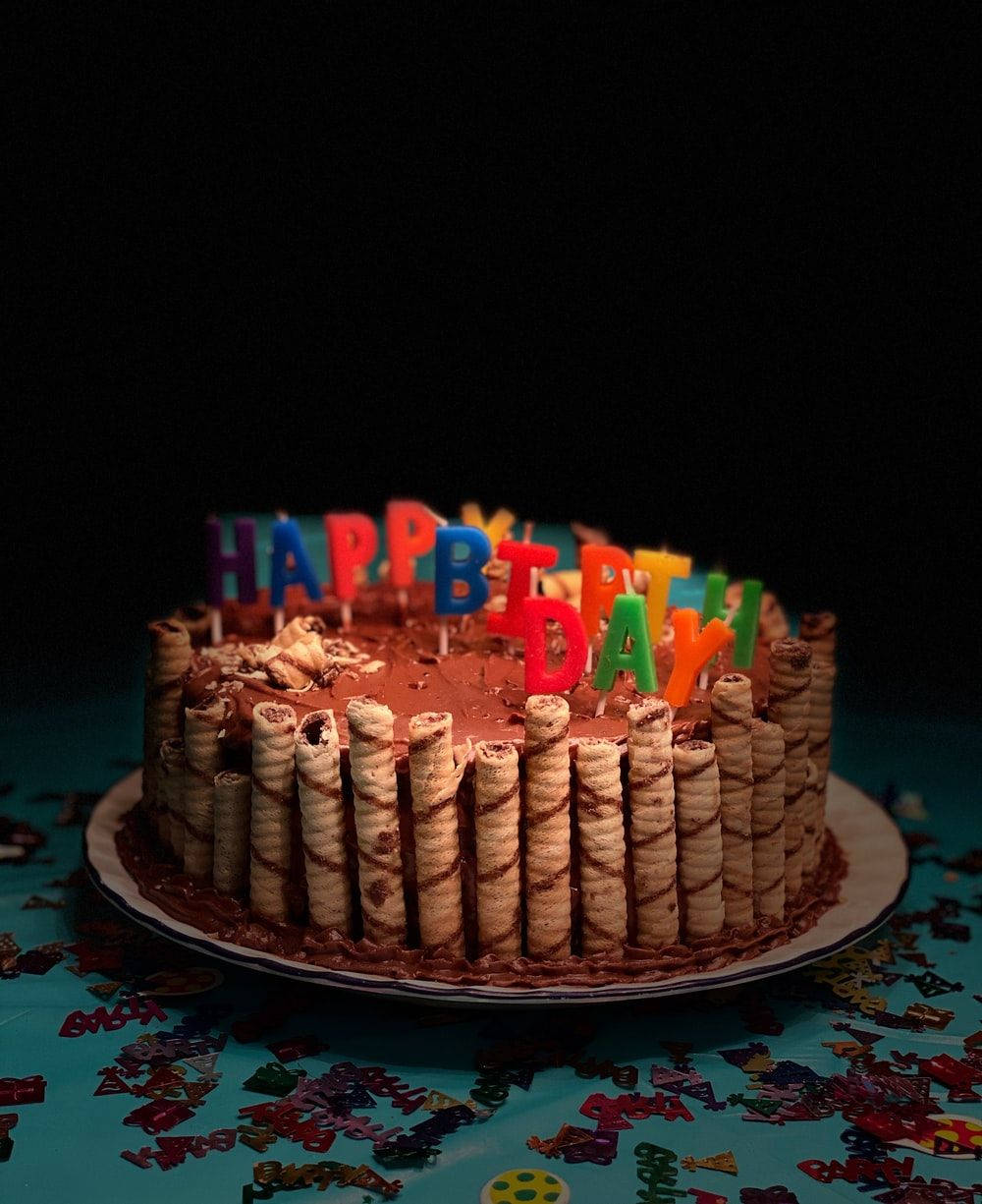 巧克力威化棒包裝生日蛋糕 壁紙 — 一個朱古力生日蛋糕，飾以彩色字母蠟燭和威化卷，放在鋪滿五彩紙屑的桌子上，在戲劇性的燈光下。