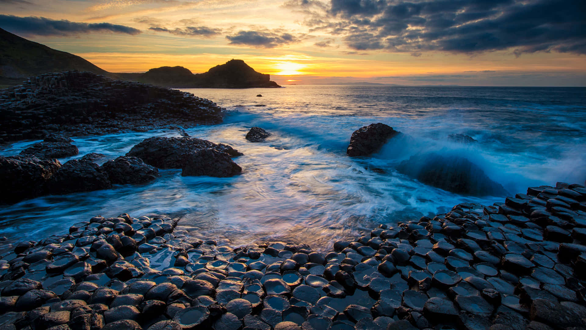 巨人堤道日落景觀 壁紙 — 夕陽將天空染成橙色，海浪拍打著崎嶇海岸場景中的玄武岩柱。