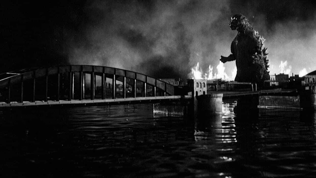 Godzilla 1954 桌布 壁紙 — 巨型怪獸哥斯拉夜晚站在燃燒的橋上，火焰和煙霧在淹沒的城市景觀上升起。