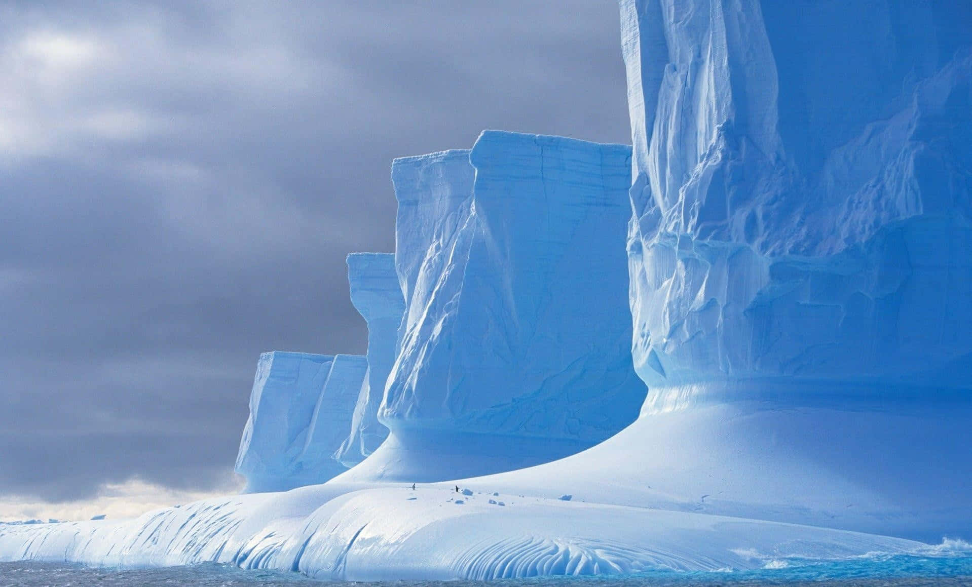 Antarctica 桌布 壁紙 — 巨大的藍色冰山從冰冷的海水中升起，天空多雲，雪地上可見小企鵝。