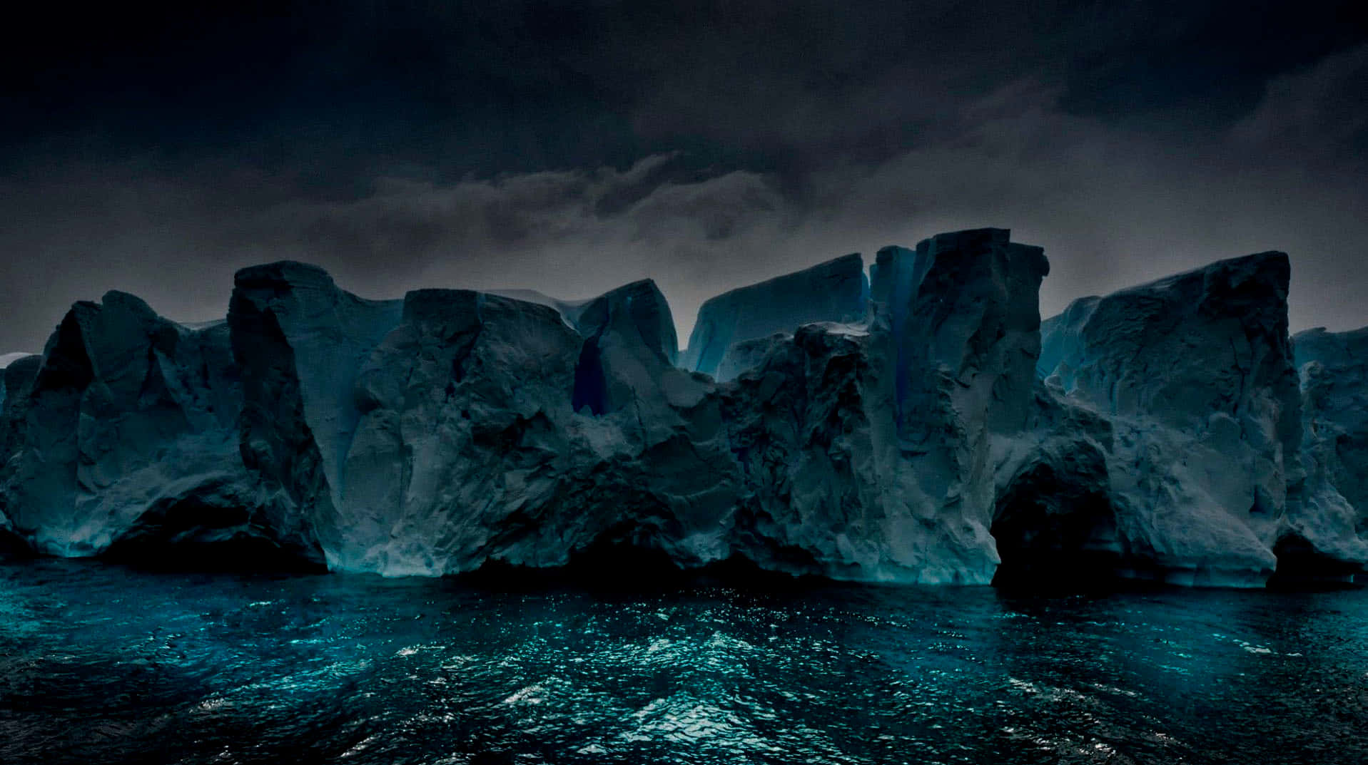 Antarctica 桌布 壁紙 — 巨大的鋸齒狀冰山從波濤洶湧的藍綠色海水中升起，上方是風暴肆虐的黑暗天空。