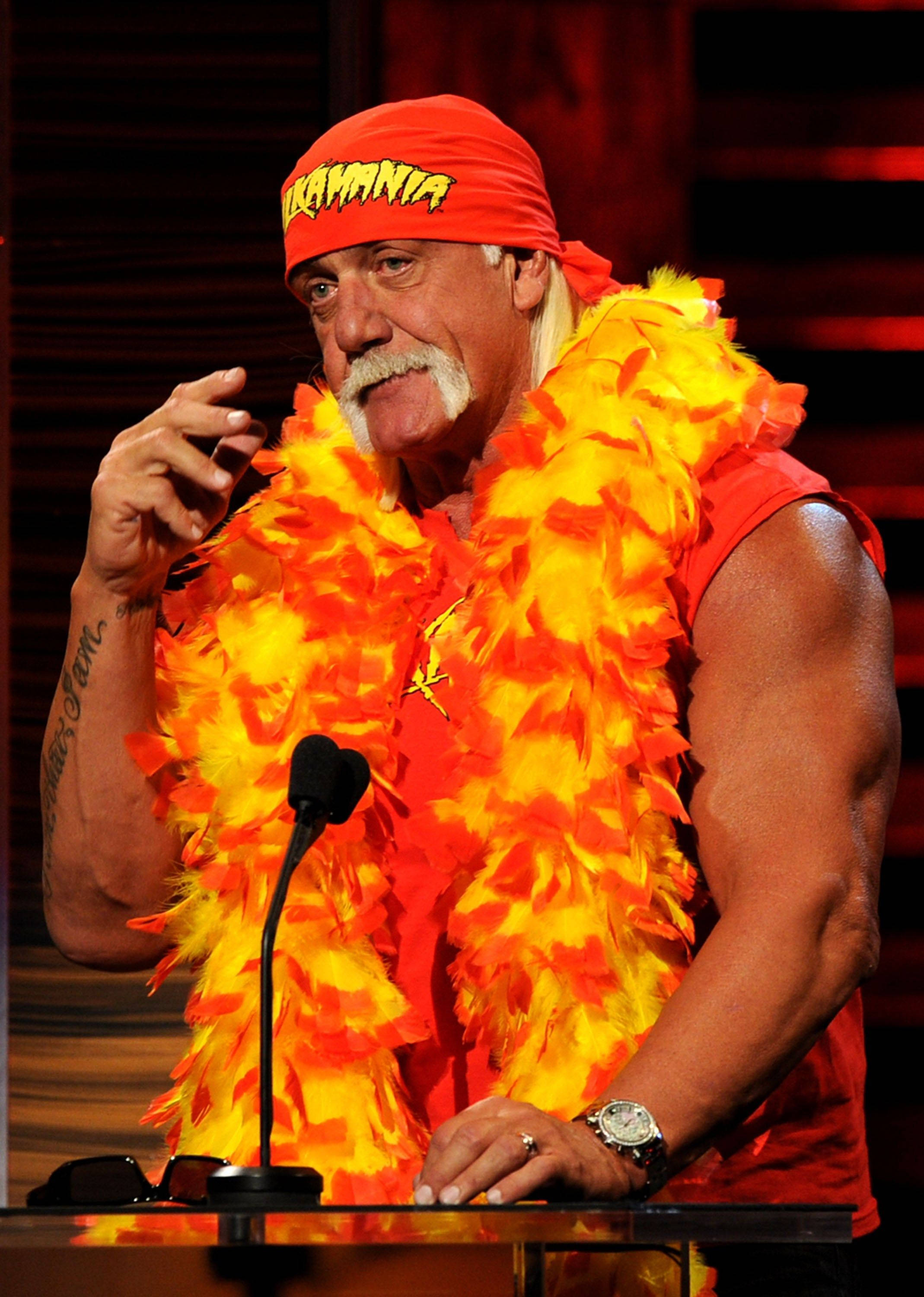巨石強森配羽毛披肩 壁紙 — Hulk Hogan 身穿標誌性的橙色和黃色羽毛服裝，站在舞台燈光下的講台上。