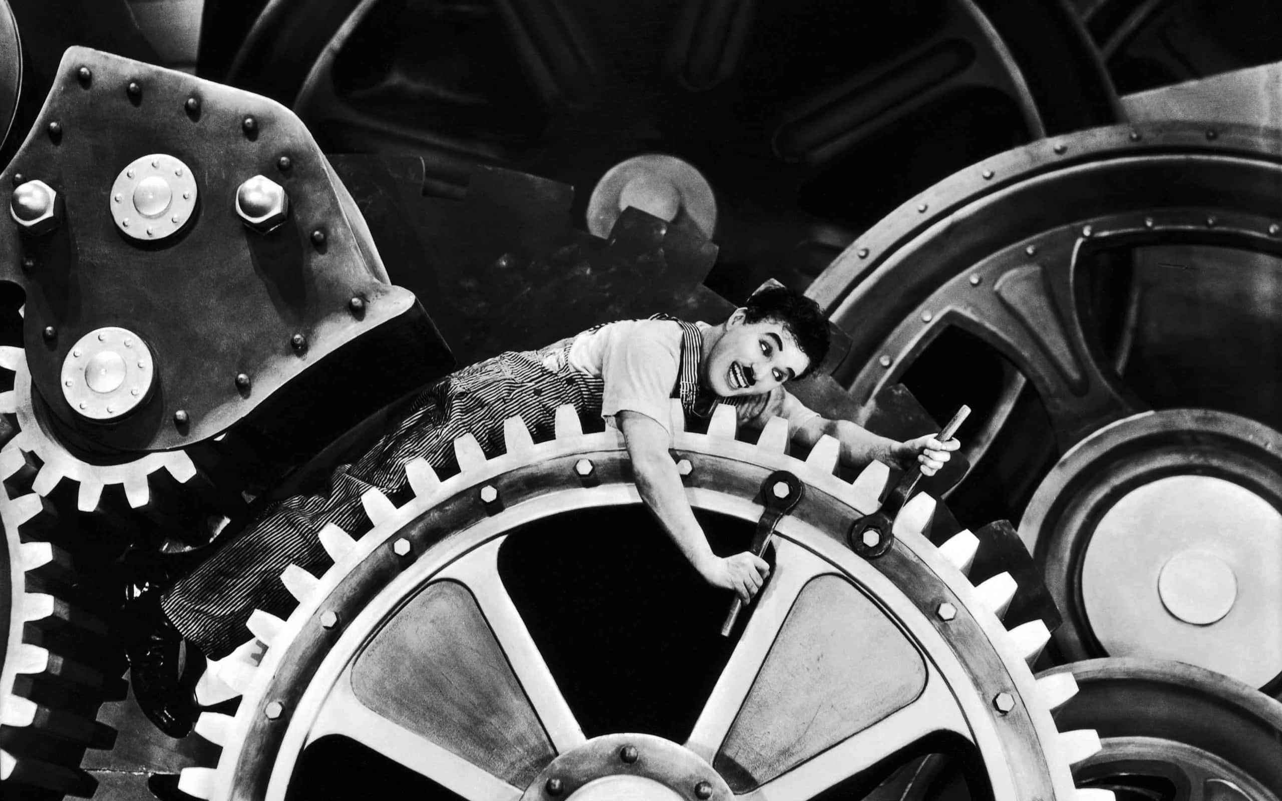 Black And White Movie 桌布 壁紙 — 差利在經典默片場景中，頑皮地咧嘴而笑，棲身於一個巨大的工業齒輪之上。