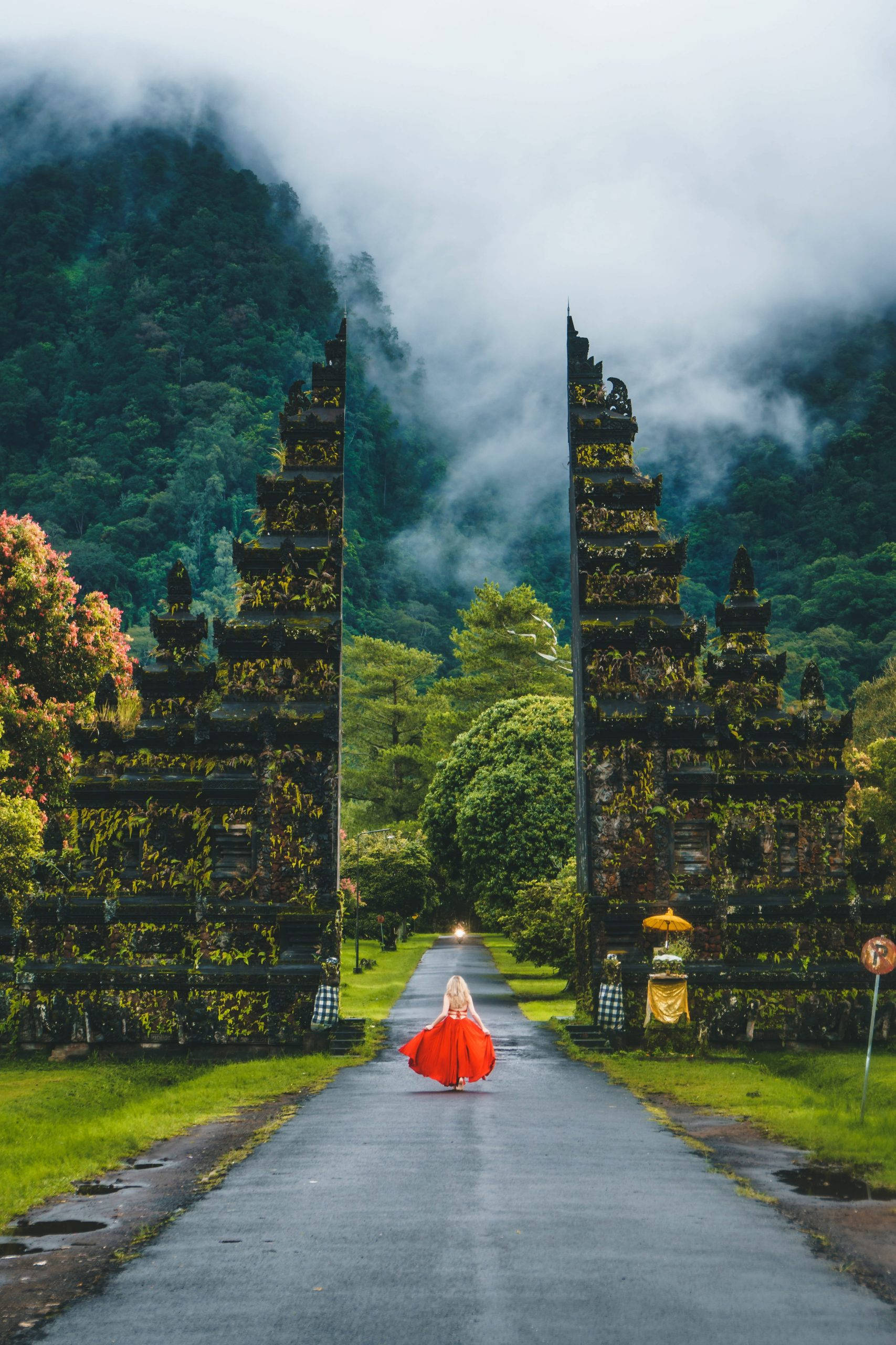 巴厘島天堂之門之旅 壁紙 — 一個孤獨的身影穿著鮮豔的紅色連衣裙，走在一條霧氣瀰漫的寺廟小徑上，兩旁是古老的石門和鬱鬱蔥蔥的綠色山脈。