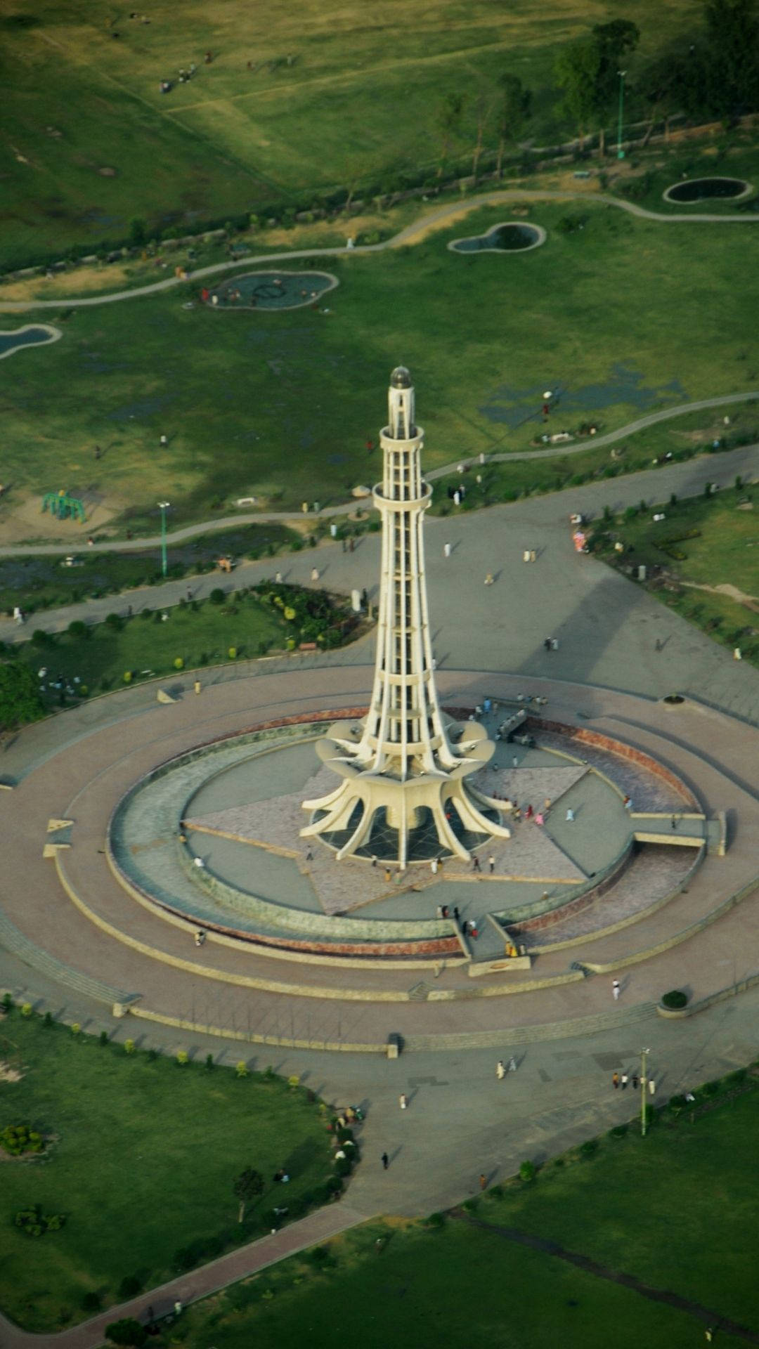 巴基斯坦 Minar-E 空拍視角 壁紙 — 高空俯瞰一座高聳的白色塔樓紀念碑，周圍環繞著圓形步道和鬱鬱蔥蔥的公園綠地，散落著遊客。