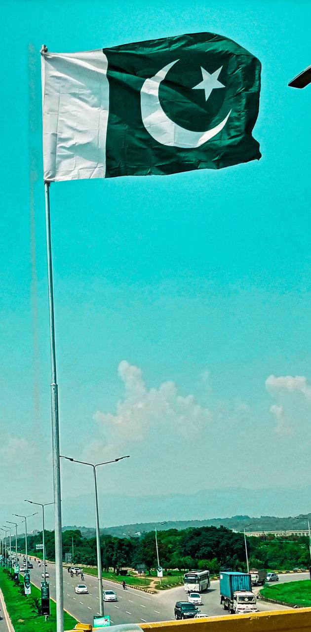 巴基斯坦國旗懸掛於旗杆 壁紙 — 巴基斯坦國旗在高高的旗桿頂端自豪地飄揚，背景是充滿活力的藍綠色天空，下方是繁忙的高速公路和遠處的山丘。