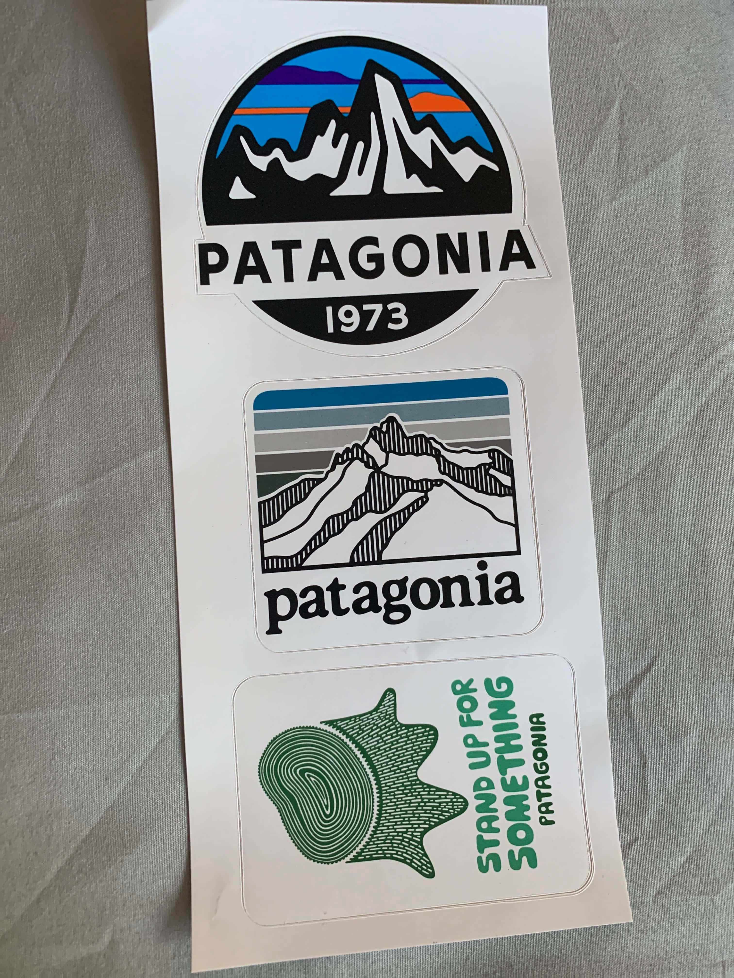 巴塔哥尼亞之美 壁紙 — 三張Patagonia品牌貼紙，上面有山脈標誌和樹木年輪設計，放在白色紙張上，背景為灰色布料。