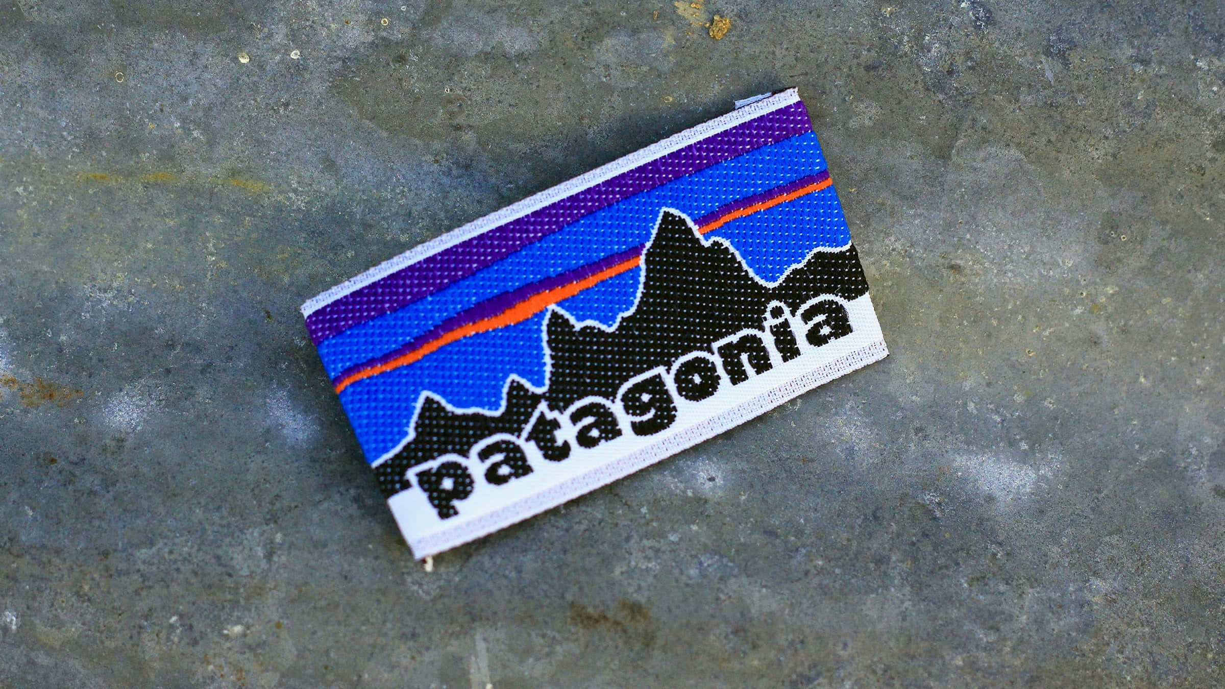 巴塔哥尼亞壯麗地平線 壁紙 — 一個帶有山脈標誌的Patagonia編織補丁放在有紋理的灰色混凝土表面上。
