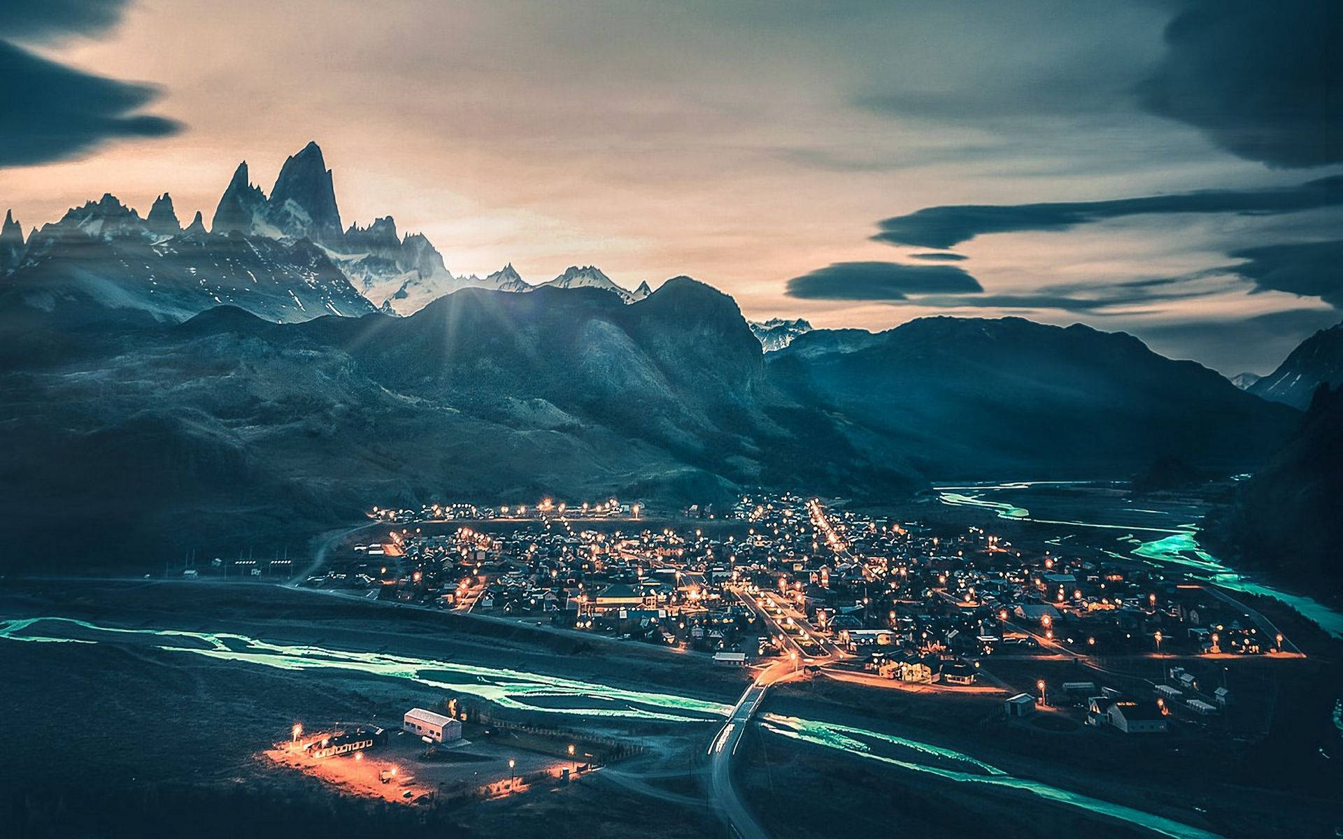 巴塔哥尼亞山腳下小鎮夜景 壁紙 — 在鋸齒狀的雪山峰頂和陰沉天空下，一座山城在黃昏時分閃爍著燈光的鳥瞰圖。