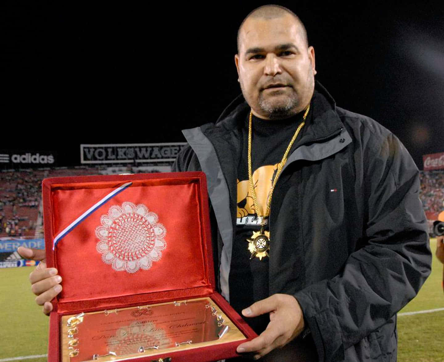 巴拉圭傳奇球員José Luis Chilavert手持阿爾塞尼奧·埃里科獎章 壁紙 — 一名身穿黑色外套的男子在夜晚的體育場上，手持一個打開的紅色天鵝絨盒子，展示獎牌和證書。