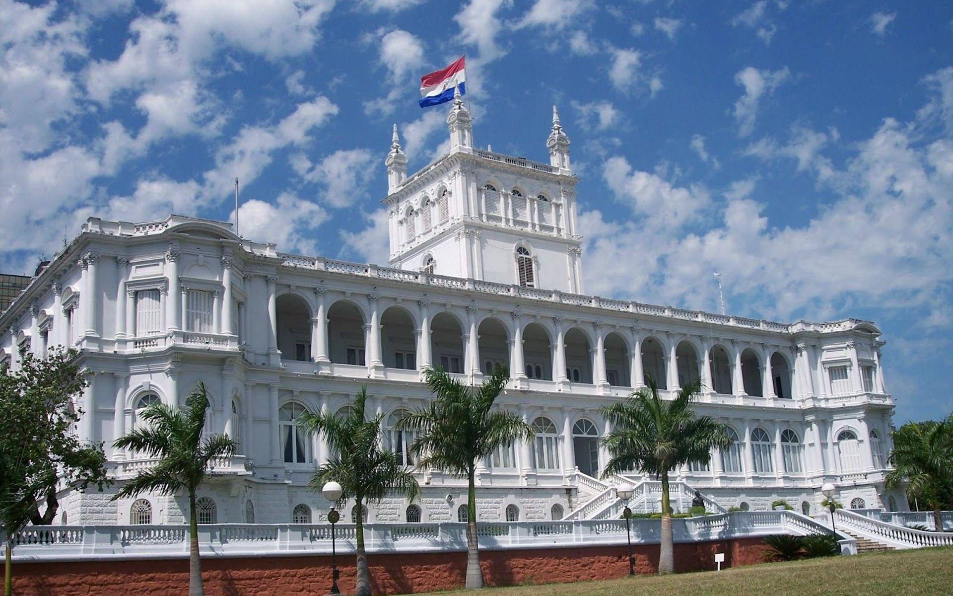 巴拉圭優雅建築 壁紙 — 一座宏偉的白色殖民風格建築，擁有拱形陽台和頂部的旗幟，周圍環繞著棕櫚樹，背景是明亮的藍天和蓬鬆的白雲。