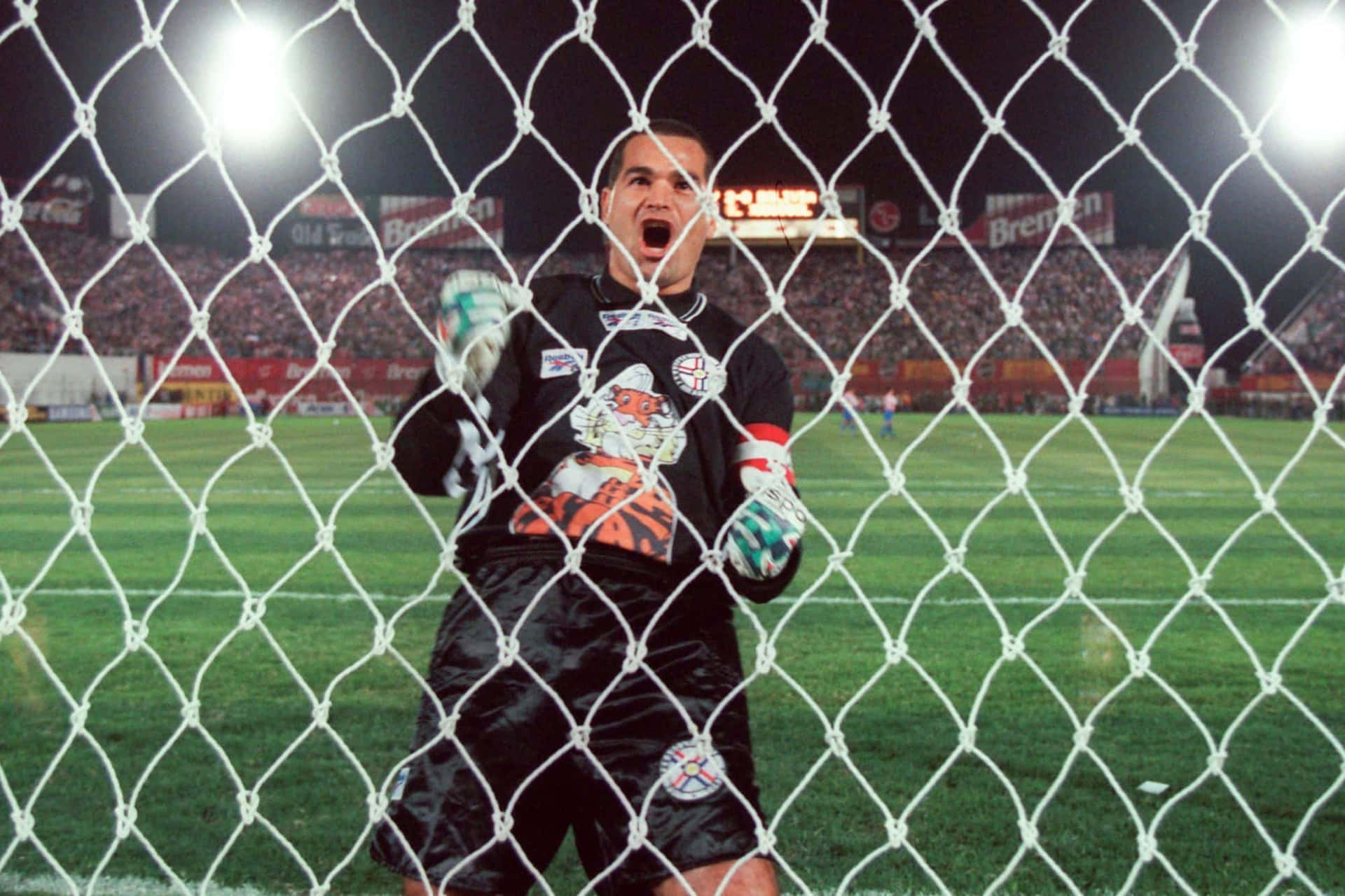 巴拉圭國家隊成員José Luis Chilavert於對陣玻利維亞時慶祝進球 壁紙 — 一名身穿黑色裝備的足球守門員在夜間球場燈光下，穿過白色球網咆哮，身後是座無虛席的觀眾。