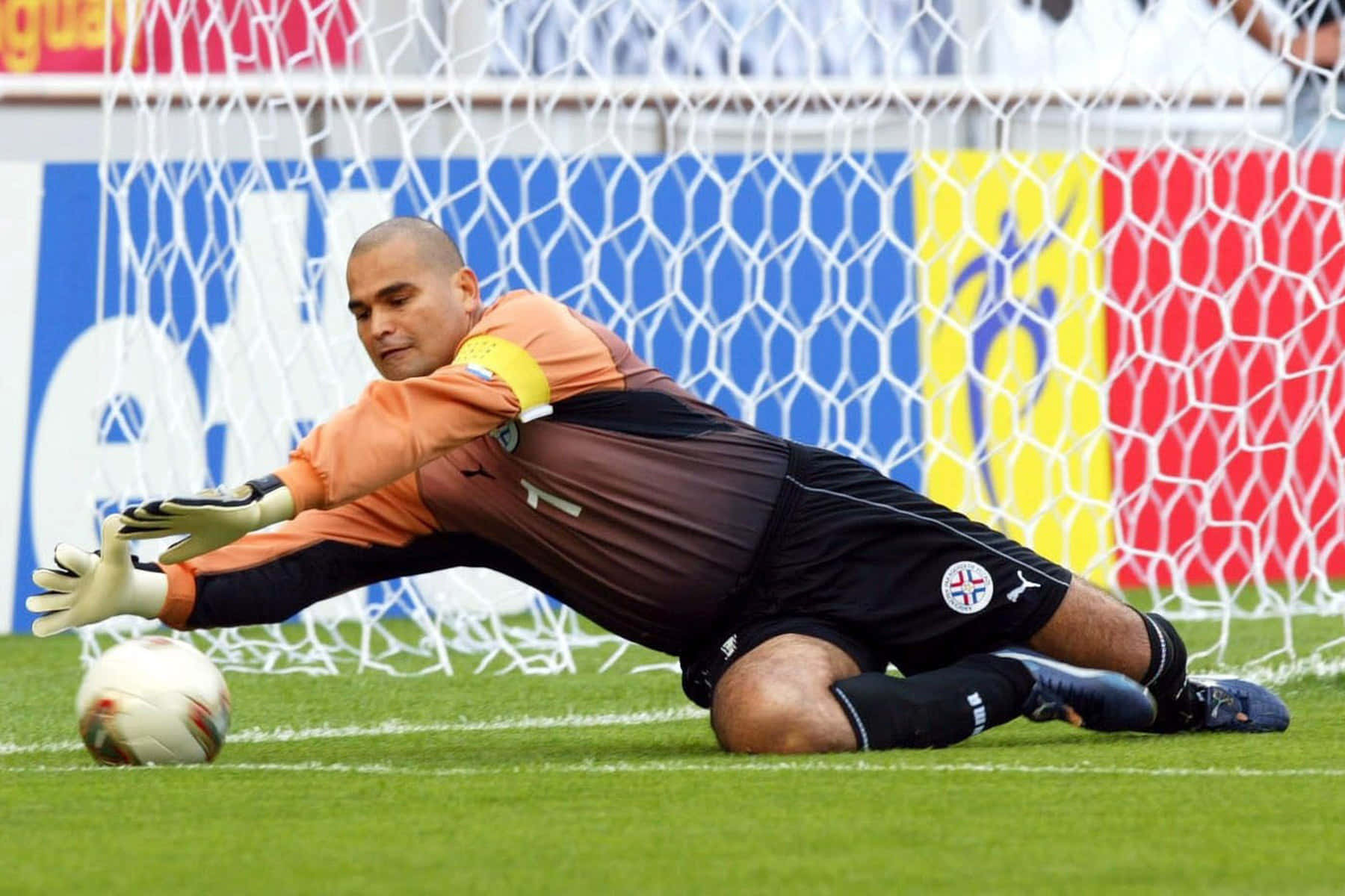 巴拉圭守門員足球員José Luis Chilavert於2002年韓國世界杯防守 壁紙 — 一位身穿橙色和黑色的足球守門員在充滿活力的綠色草地上低飛撲救，靠近色彩繽紛的球門網。
