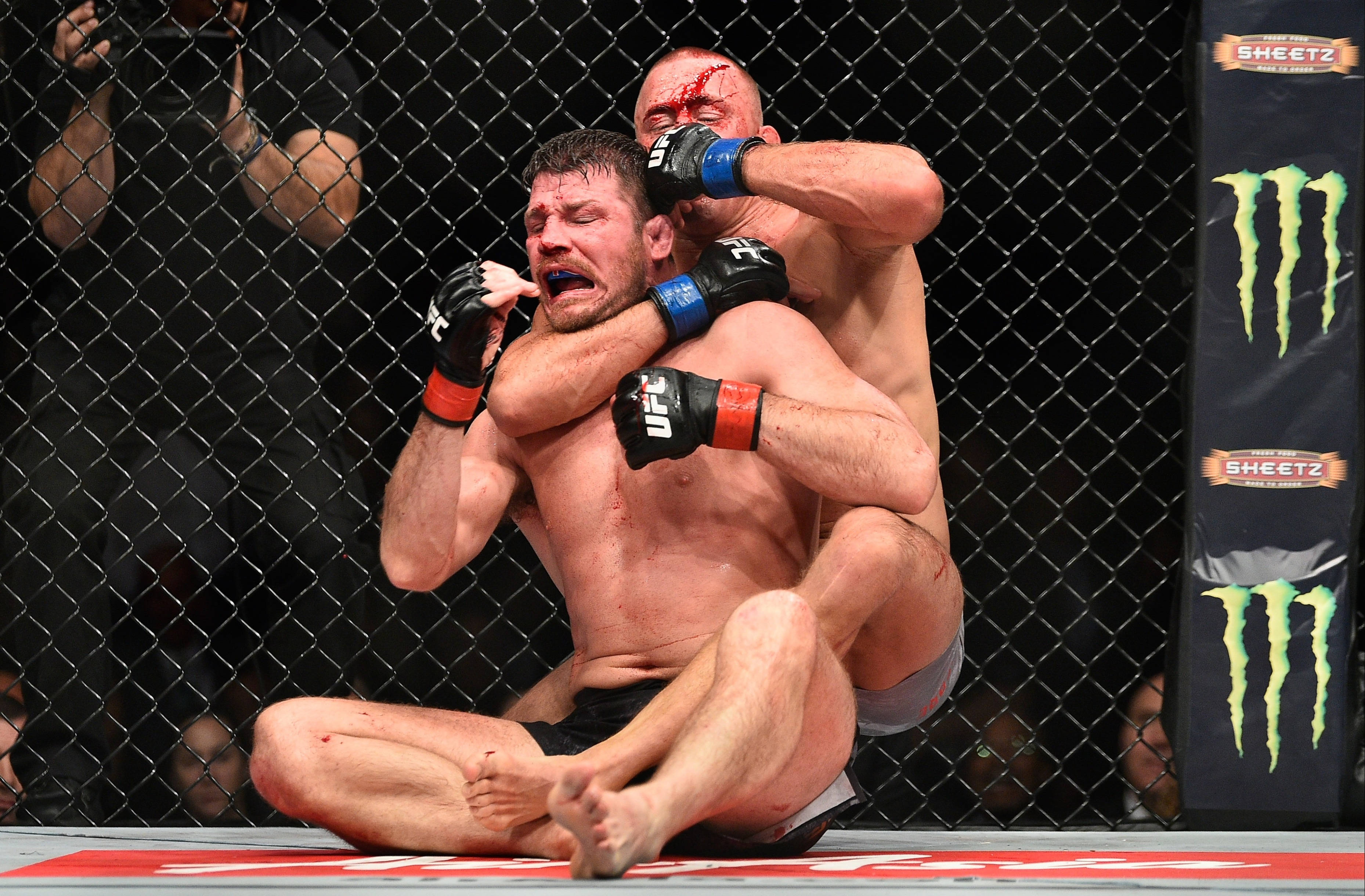 巴斯·魯滕施以頭鎖 壁紙 — 兩名血跡斑斑的MMA拳手在八角籠內激烈纏鬥，籠上有明顯品牌標誌，背景有觀眾。