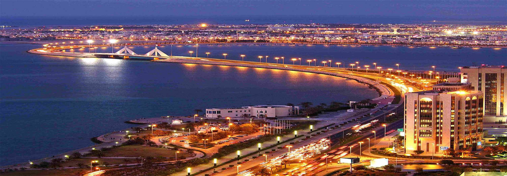 巴林夜景 壁紙 — 現代海岸城市在暮色中的驚人景色，燈光閃爍，倒映在平靜的水面上，還有一座彎曲的照明橋樑。
