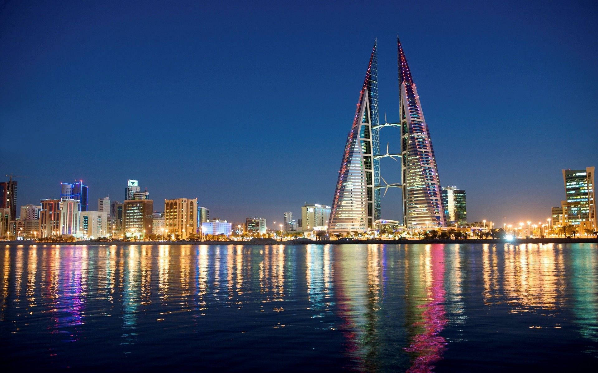 巴林夜間海景 壁紙 — 標誌性的巴林世界貿易中心塔樓在夜間閃爍著彩色燈光，倒映在平靜的水面上。