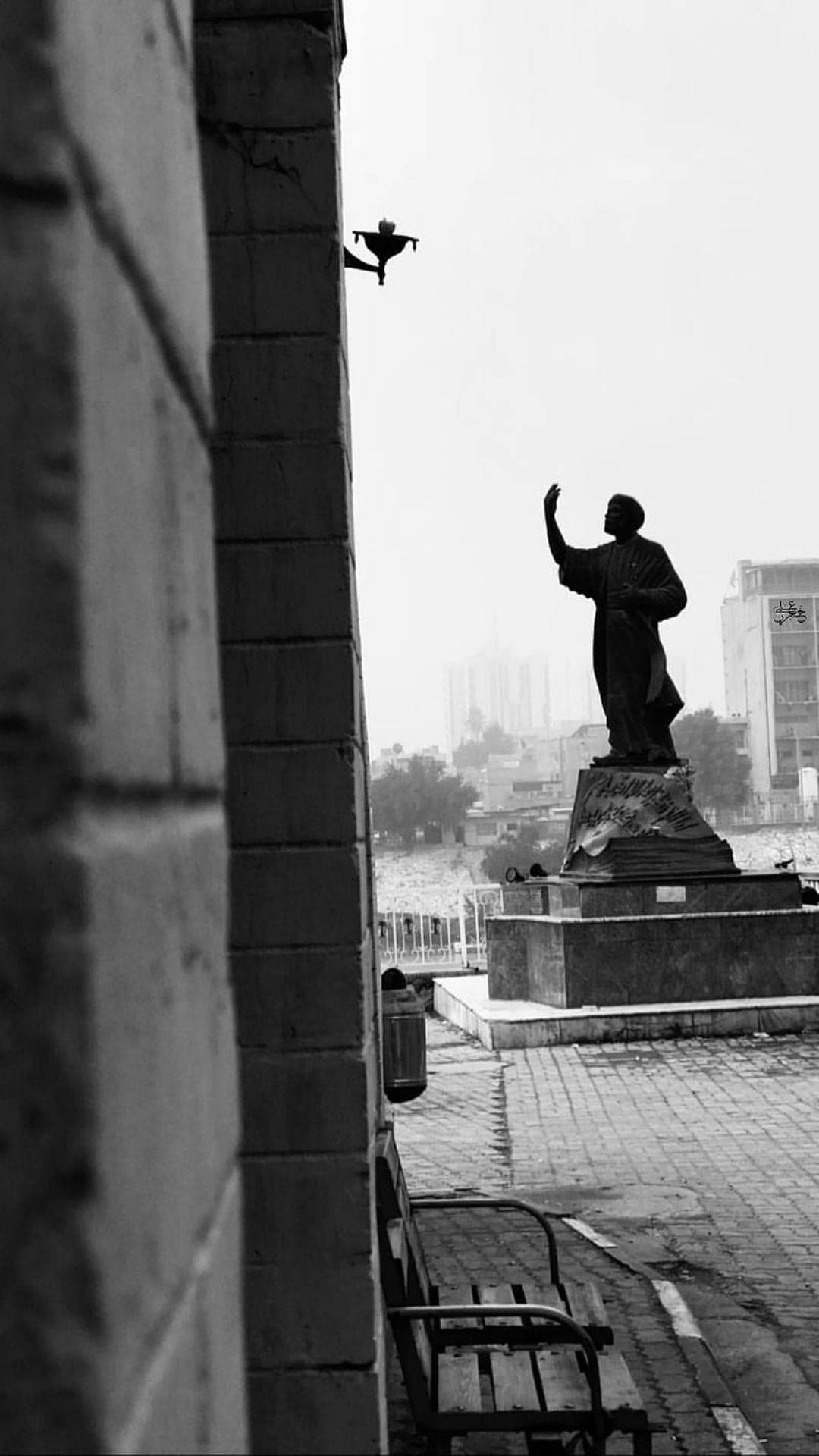 巴格達著名雕像 壁紙 — 黑白城市場景，一座舉臂雕像立在磚牆旁，一隻鳥棲息在壁架上。
