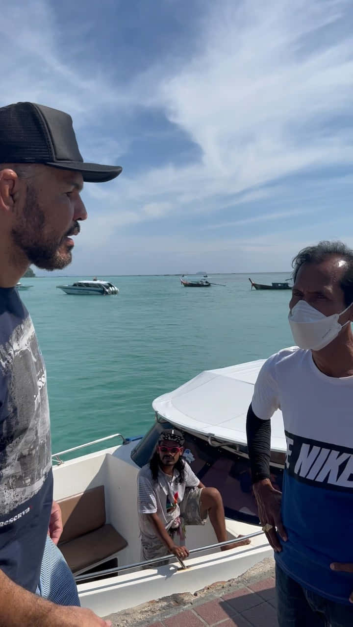 三個男人喺熱帶碼頭嘅船上，周圍有藍綠色海水同遠處嘅船。