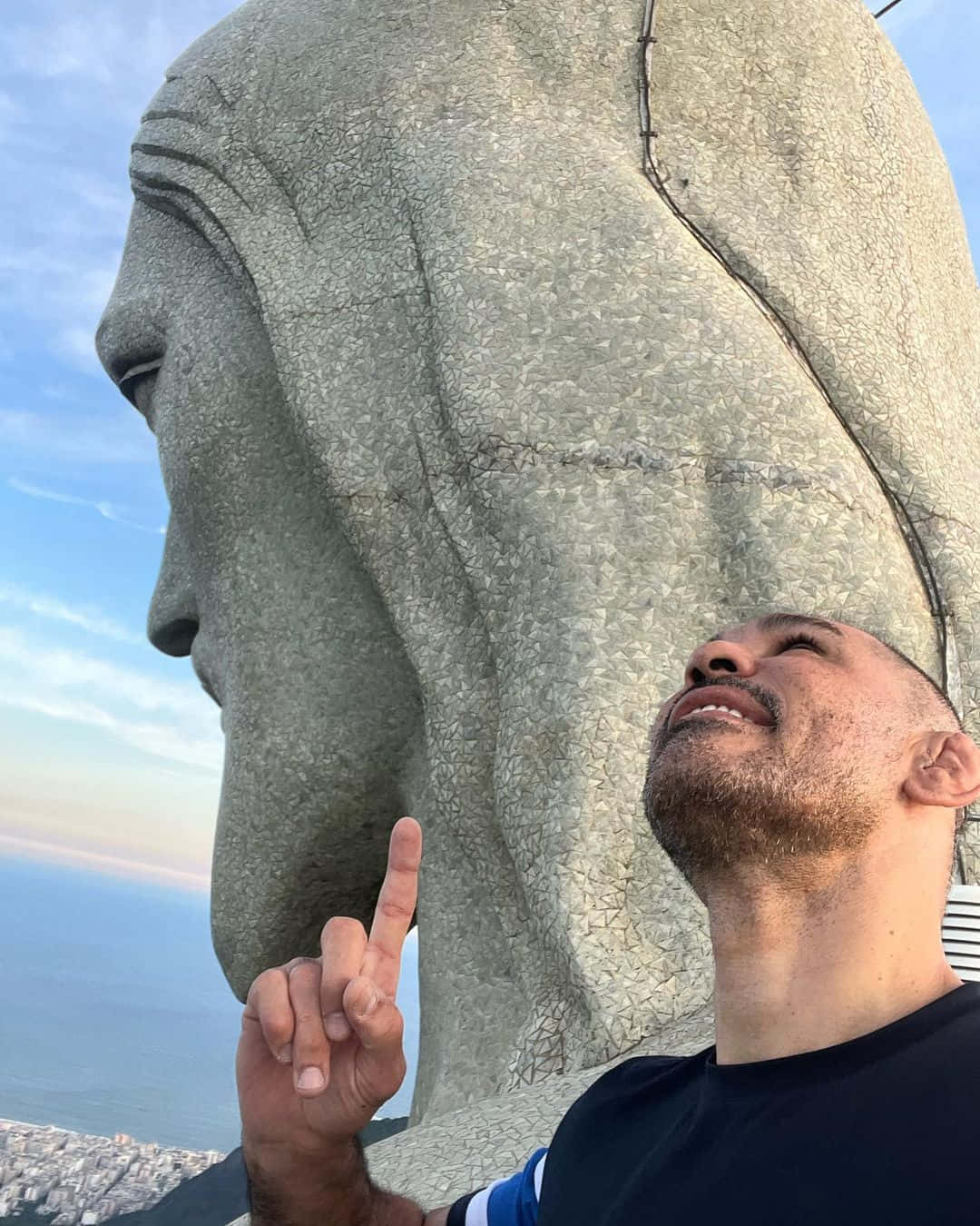 巴西MMA戰士安東尼奧·羅德里戈·諾格拉在比賽中 壁紙 — 一名男子抬頭微笑，面向巨大的基督救世主雕像，背景是清澈的藍天，下方有海洋和城市。