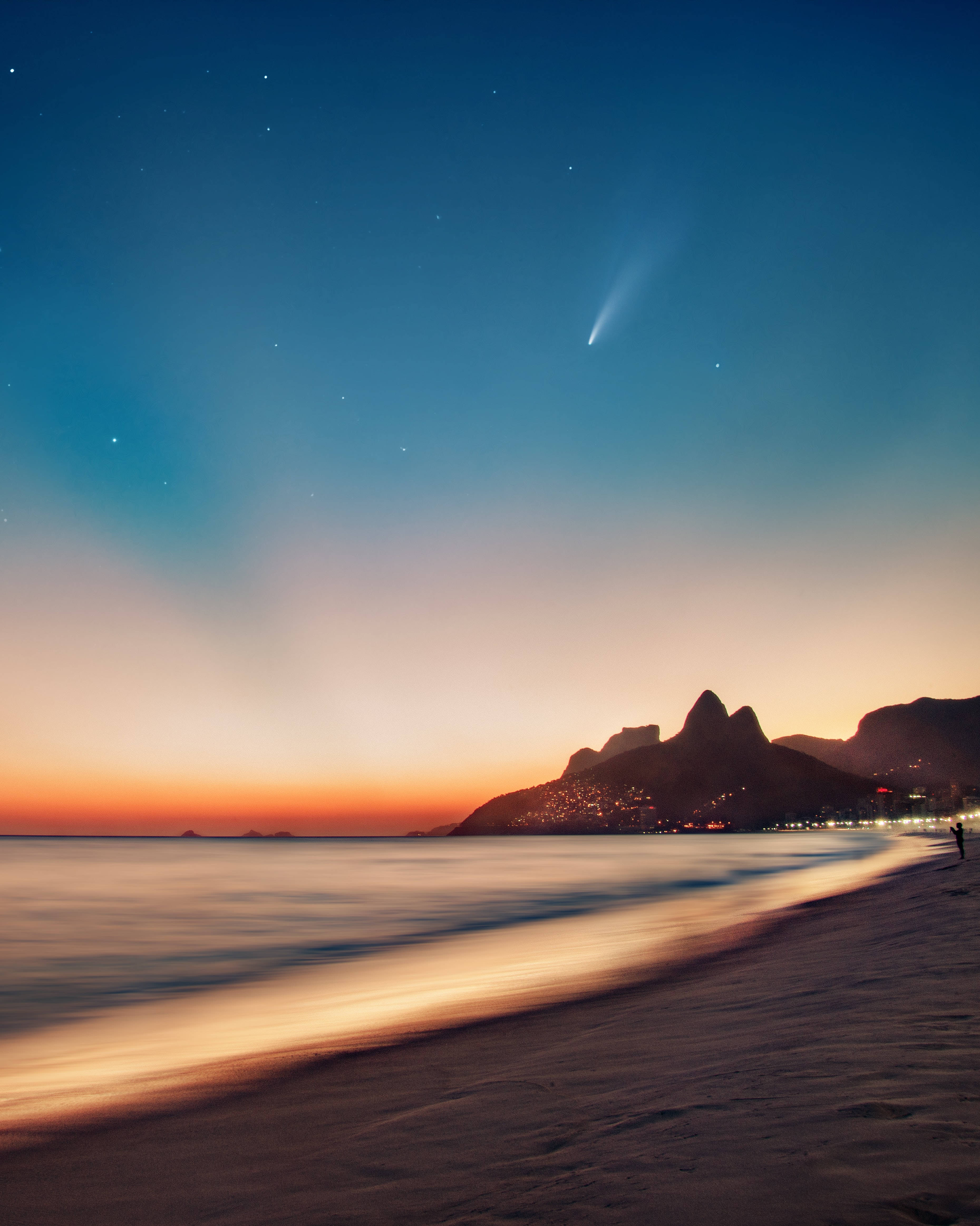 巴西伊帕內瑪海灘 壁紙 — 一顆彗星劃過暮色天空，下方是寧靜的海灘，城市燈光閃爍，山巒輪廓分明。