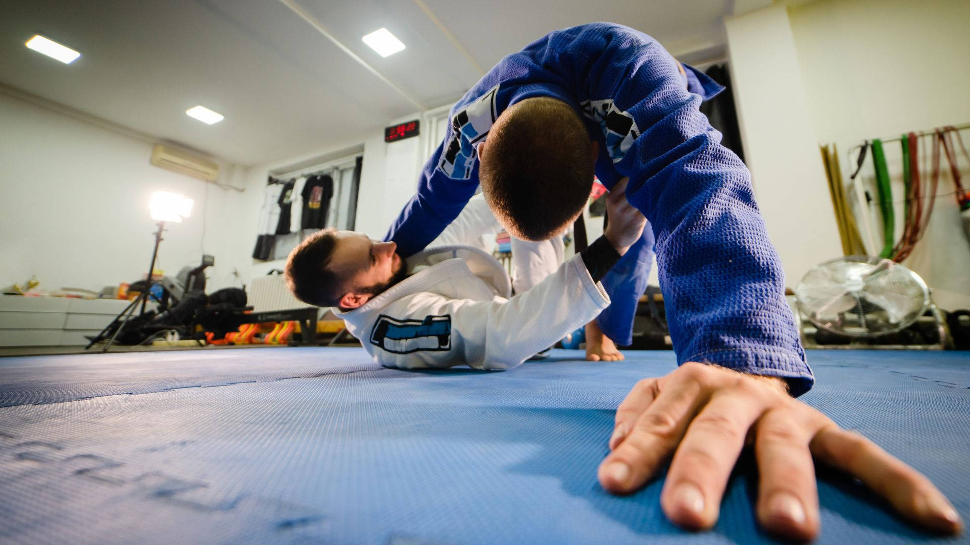 巴西柔術武術對抗動作 壁紙 — 兩名武術家在光線充足的健身房的藍色軟墊上激烈纏鬥，背景有訓練器材。