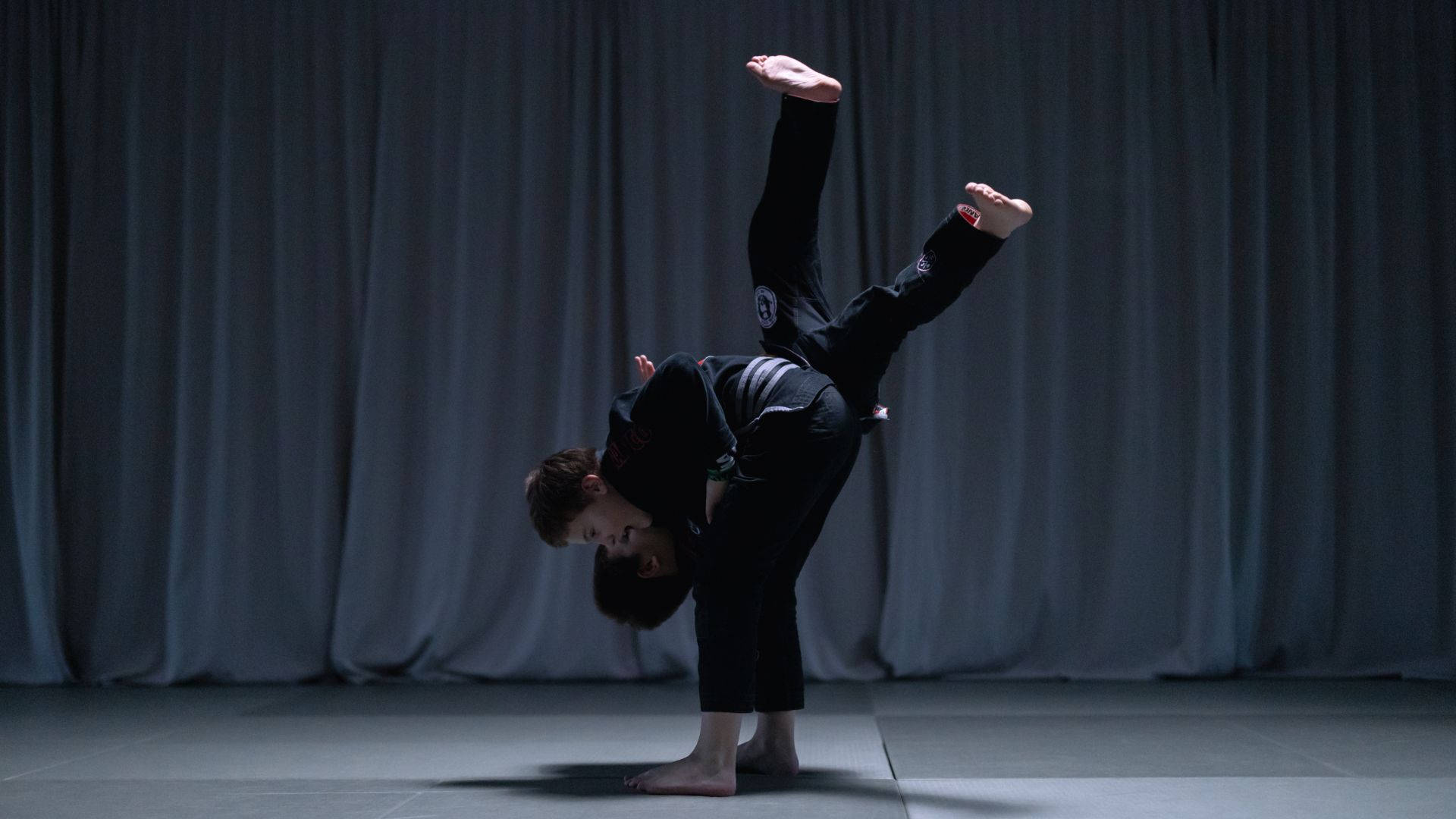 巴西柔術的力量——格鬥 Mastery 的展現 壁紙 — 一名武術家在黑暗帷幕前，於戲劇性聚光燈下在墊子上做出動態的倒立劈腿動作。
