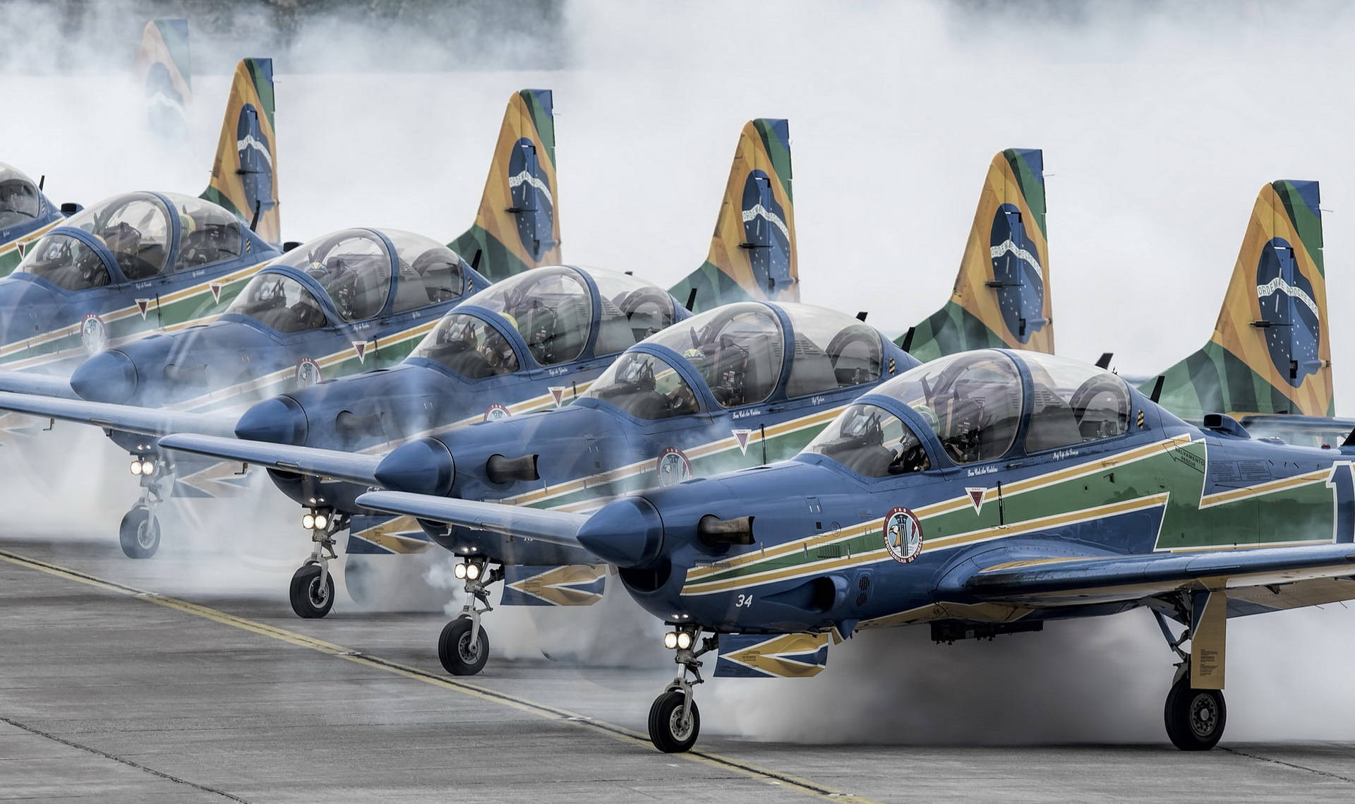 巴西空軍飛機 壁紙 — 一排巴西軍用噴射機，機尾帶有國旗，在跑道上排列，引擎噴出煙霧。